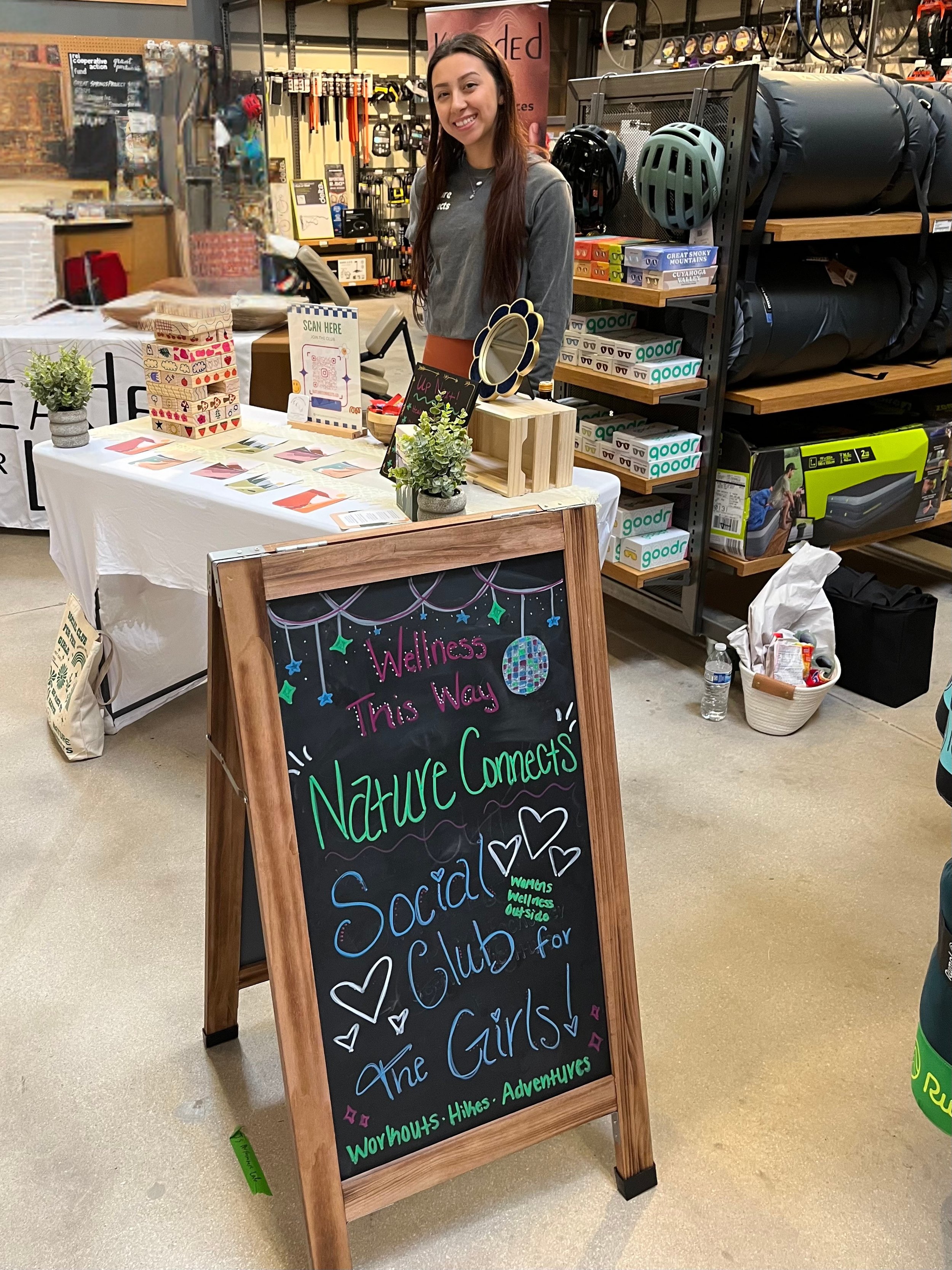A young woman with long brown hair smiling stands behind a table at an indoor market or shop. The table displays small potted plants, decorative items, and a sign instructing to scan a QR code. In front of the table is a blackboard sign with colorful handwritten text promoting a wellness and social club for girls that emphasizes nature, adventure, and outdoor activities. Shelves behind her hold bike helmets, boxes, and outdoor gear.