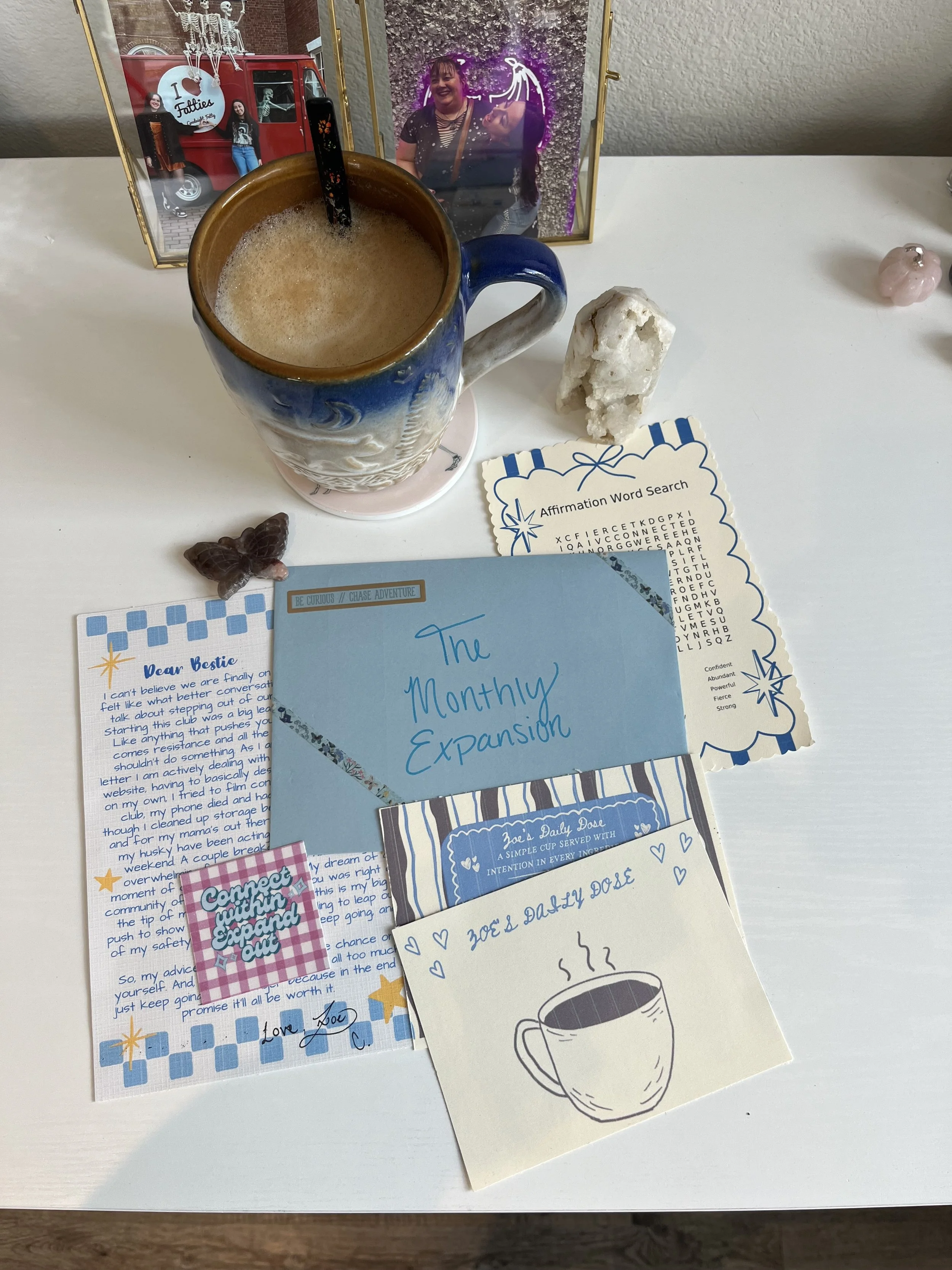 A desk decorated with a framed photo of two women smiling, a blue mug filled with a frothy beverage, a white rock, affirmation and note cards, a butterfly-shaped decoration, and some small pink objects.
