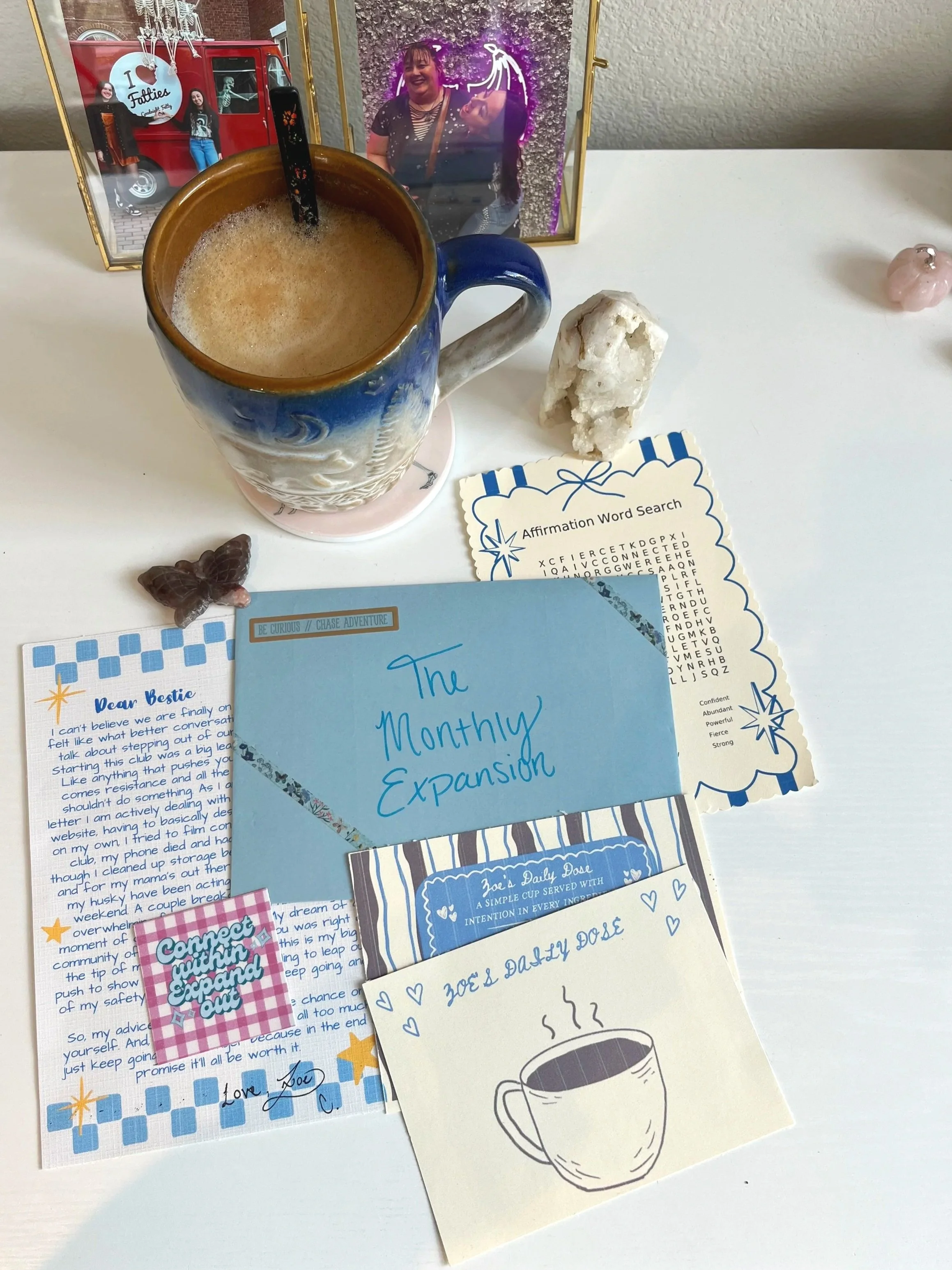 Collection of various postcards, a mug filled with a frothy beverage, a crystal large white rock, and framed photos of women, one of whom is smiling, and another of two women standing in front of a red ice cream truck with a skeleton decoration. There is also a small pink pig figurine and a coffee drawing on one of the postcards.