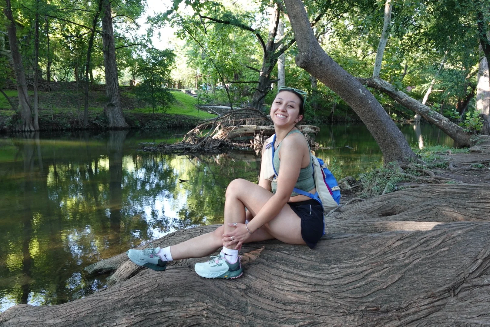 Young woman sitting on a large tree trunk by a calm river in a wooded park, smiling, with sunglasses on her head, wearing a tank top, shorts, and sneakers, with a blue backpack.