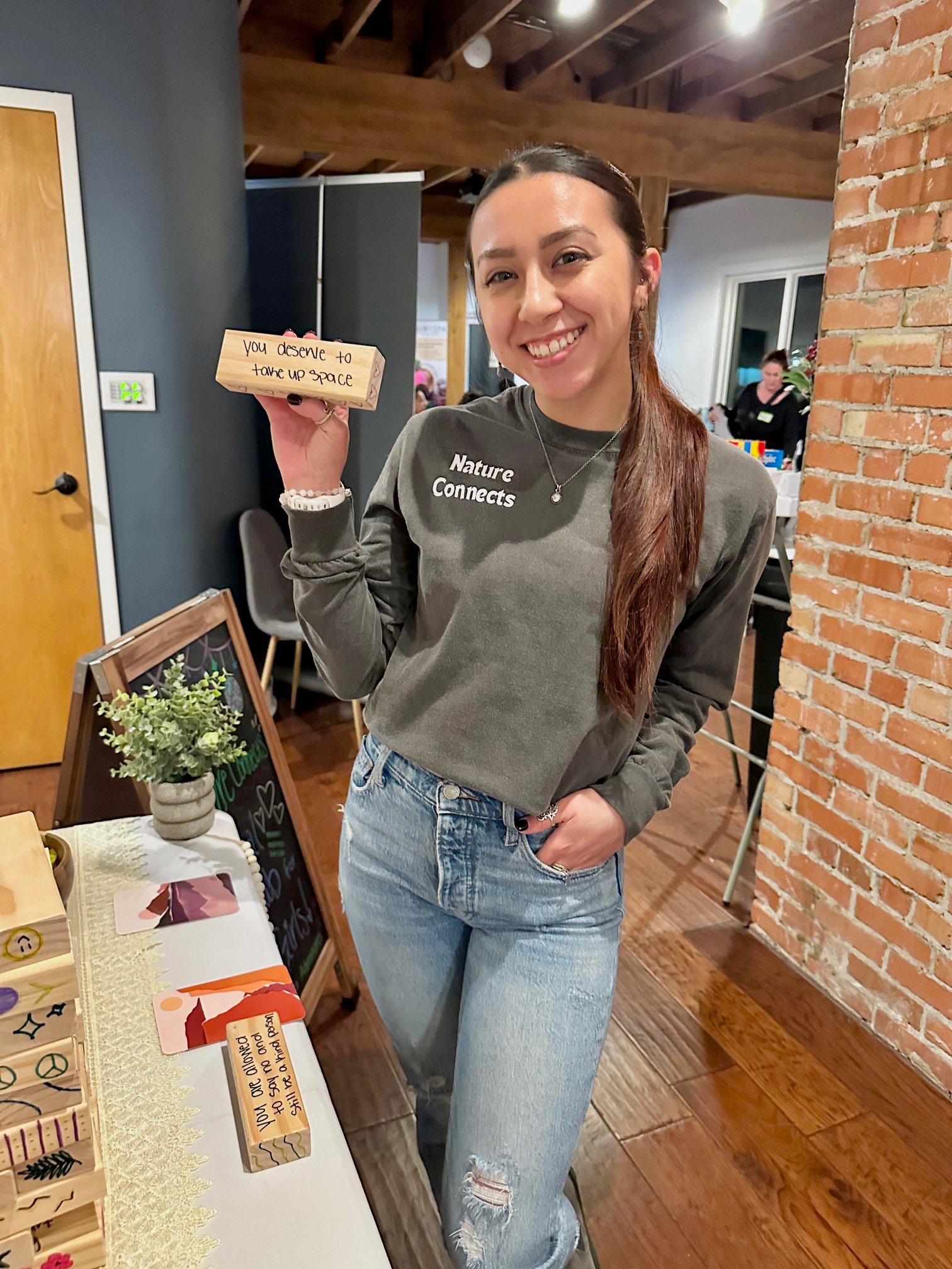 A woman holding a small wooden block with an inspirational message, standing next to a table with plants and decorative items, in a cozy indoor setting with exposed brick wall and wooden beams.