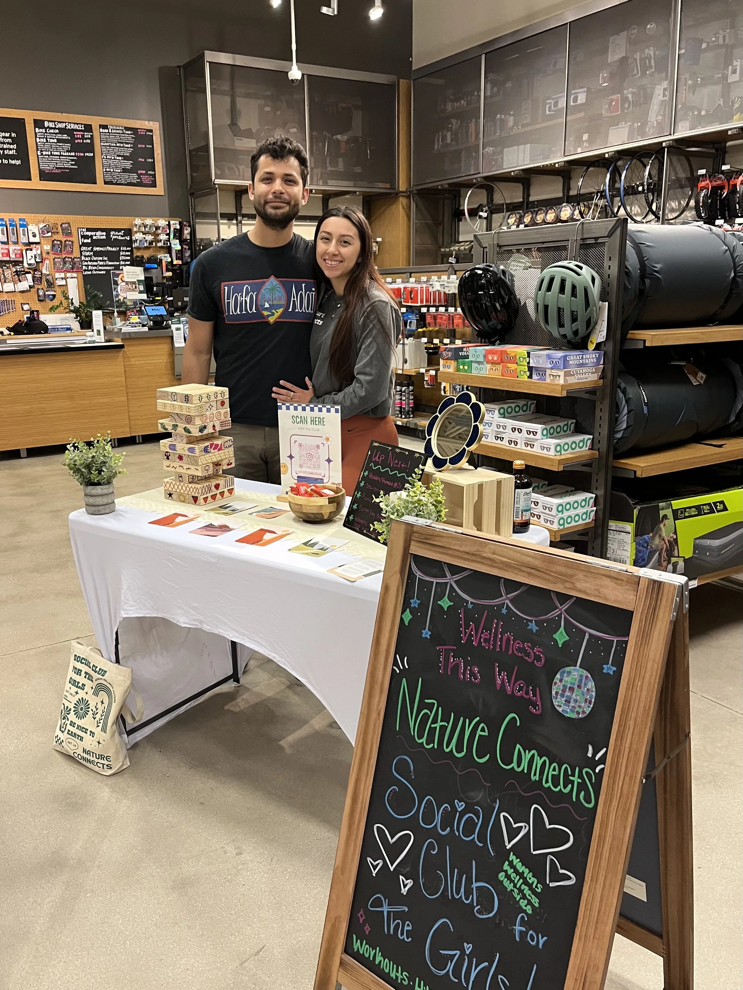 A young man and woman standing behind a table at a retail store, smiling for the camera. The table displays small decorative wooden blocks, a sign inviting people to scan for membership, and small plants. Behind them, shelves hold bicycle helmets, and a chalkboard sign features colorful writing about a social club for girls.