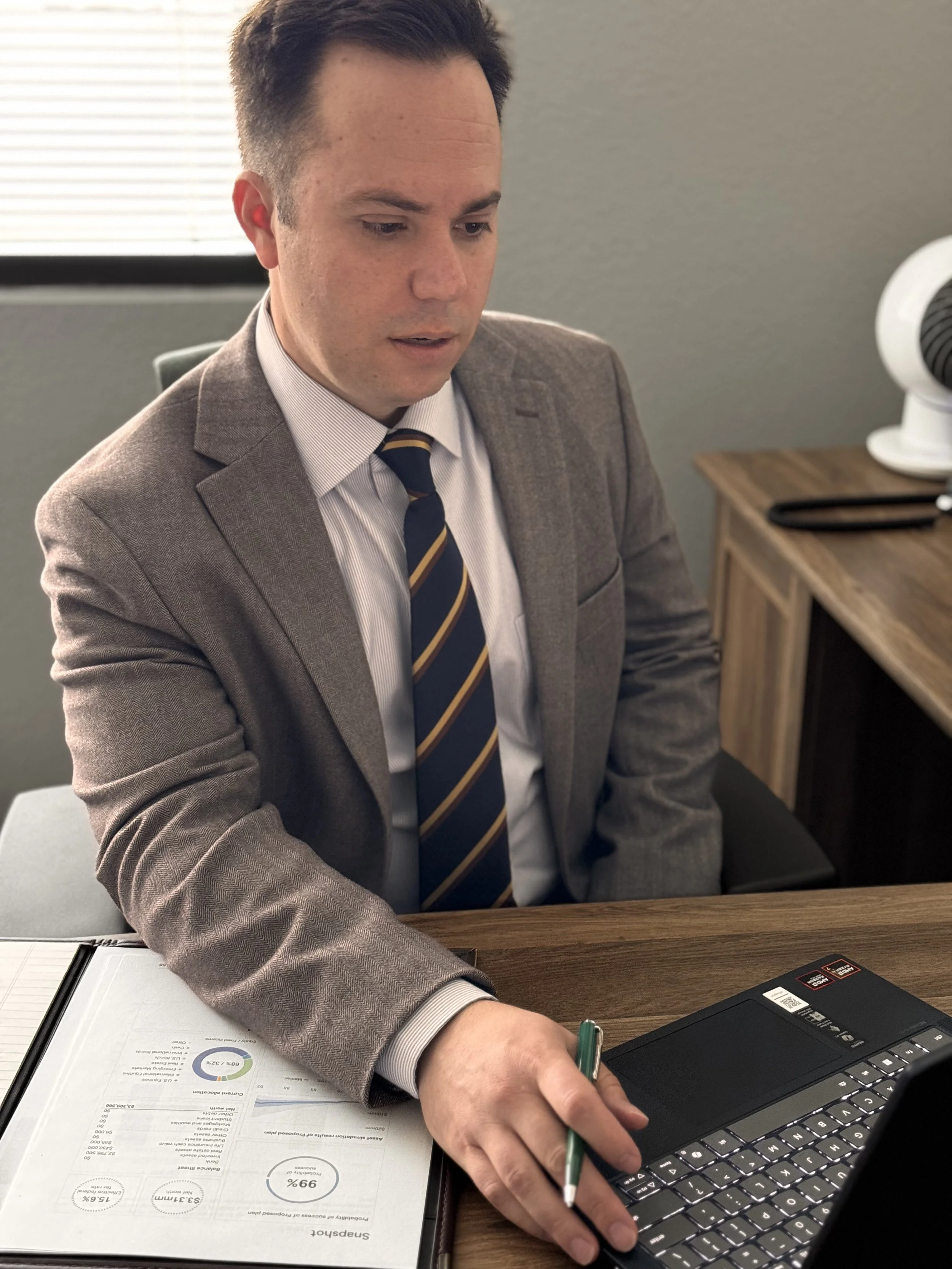 A man in a brown suit and striped tie working at a desk, using a laptop and writing on documents with charts and graphs, in an office setting.