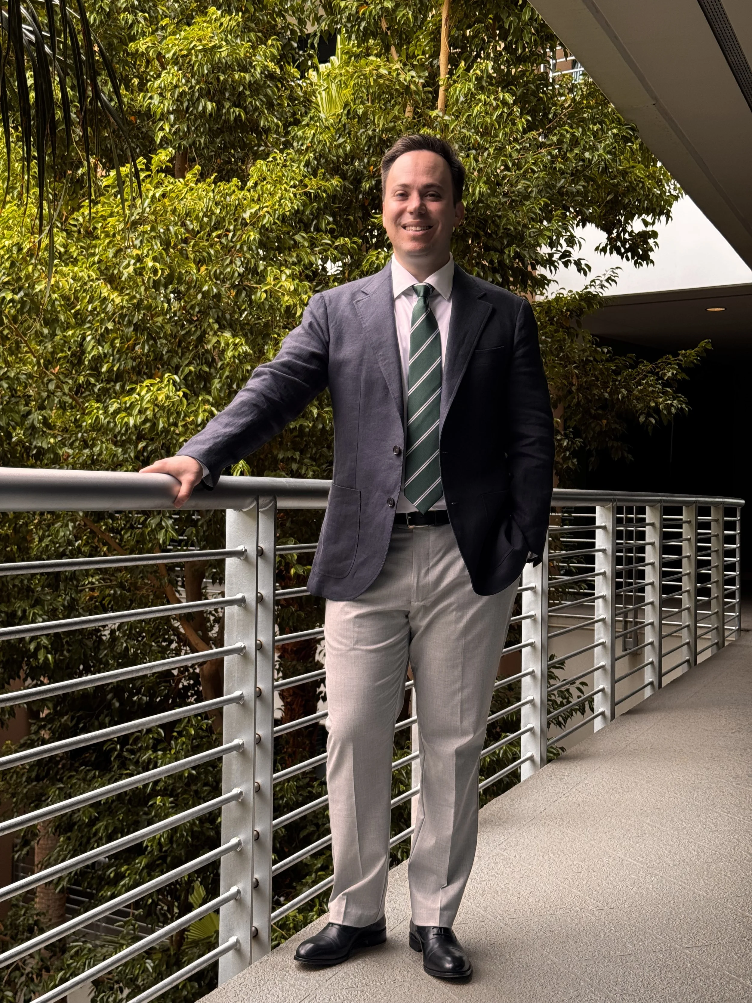 A man in a suit standing on a balcony with metal railings and lush green trees in the background.