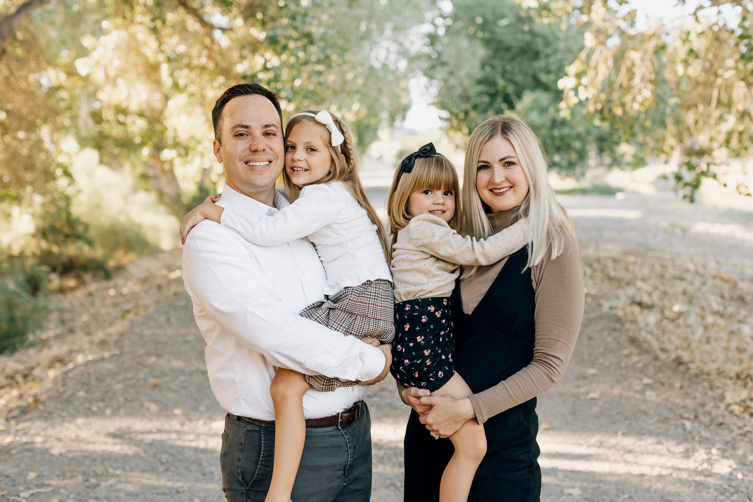 A happy family of four smiling outdoors in a park with trees and sunlight, with a father holding a young girl, mother holding her younger daughter, all close together.