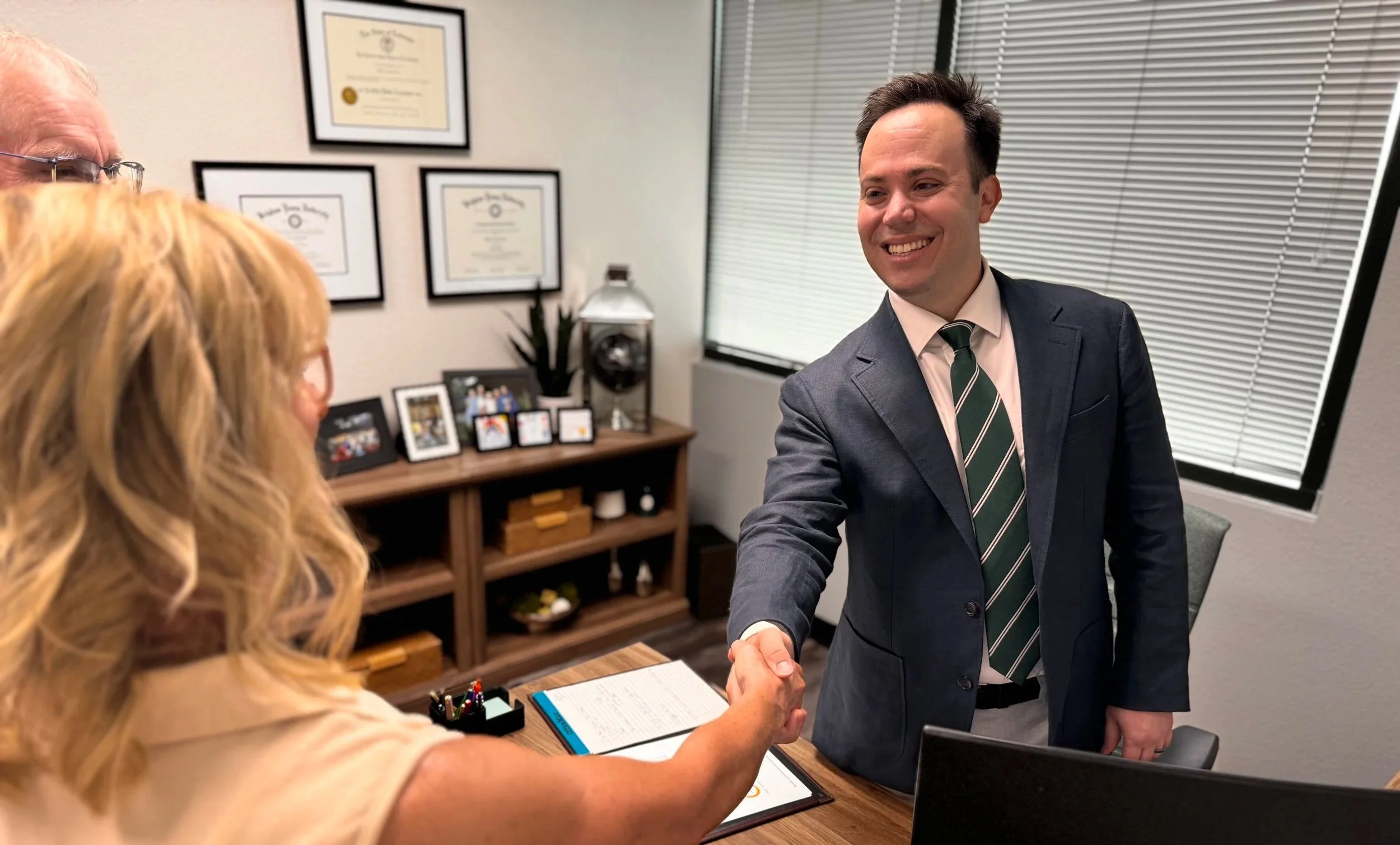 Advisor in a suit and tie shaking hands with a woman, with her partner partially visible in the foreground. The setting is an office with framed certificates on the wall, a desk with a pen holder, and items displayed on a shelf.