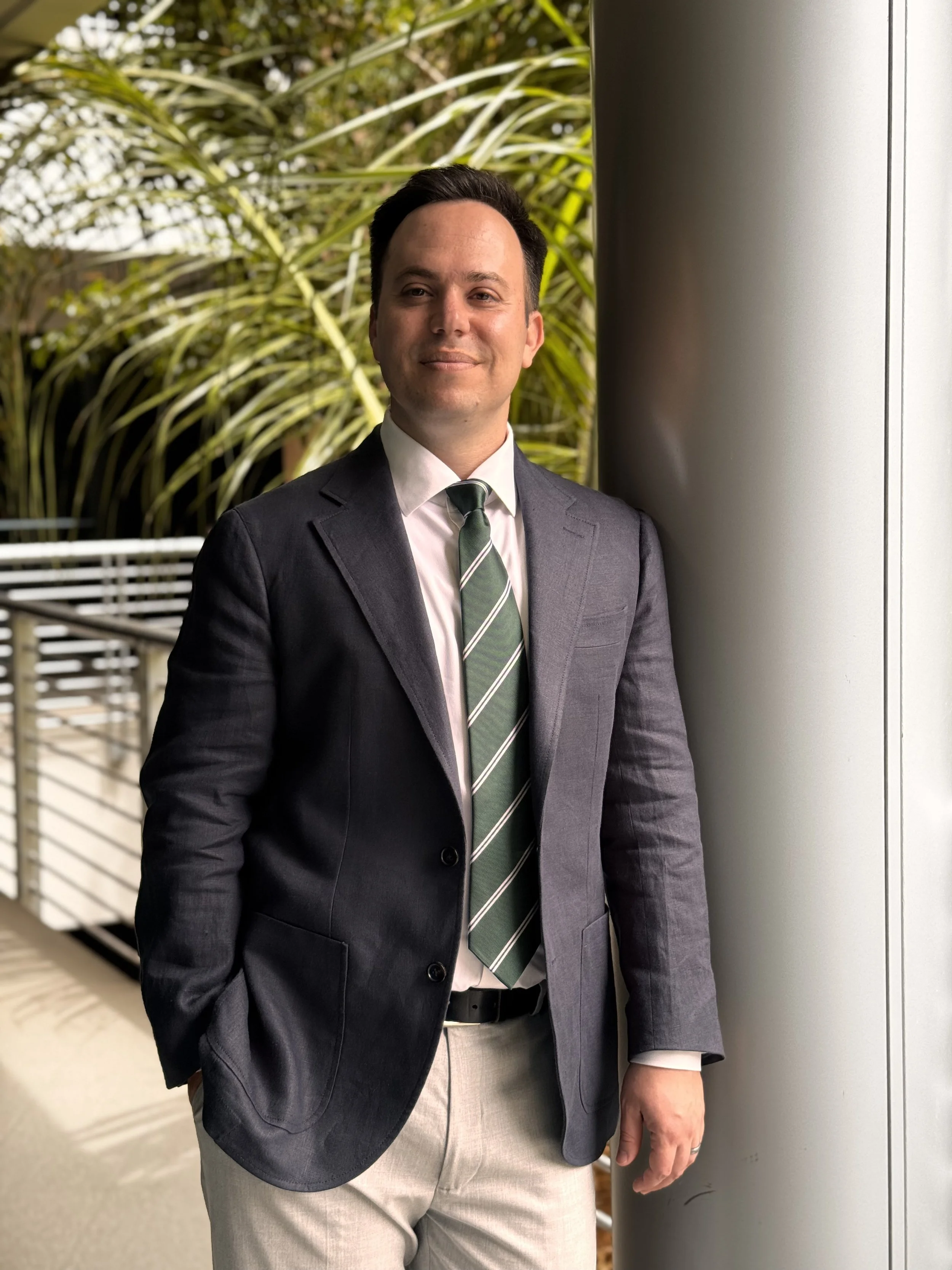 A man in a dark suit, white shirt, and green striped tie standing next to a metallic column with greenery in the background.