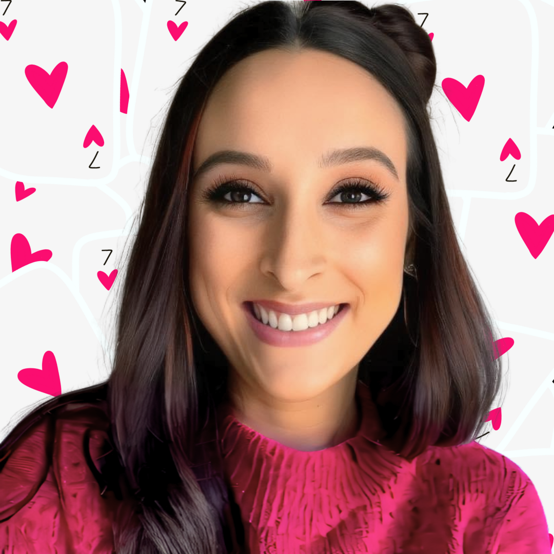 A woman with dark hair smiling, wearing a pink patterned top, with a background of pink hearts and white playing card suits.