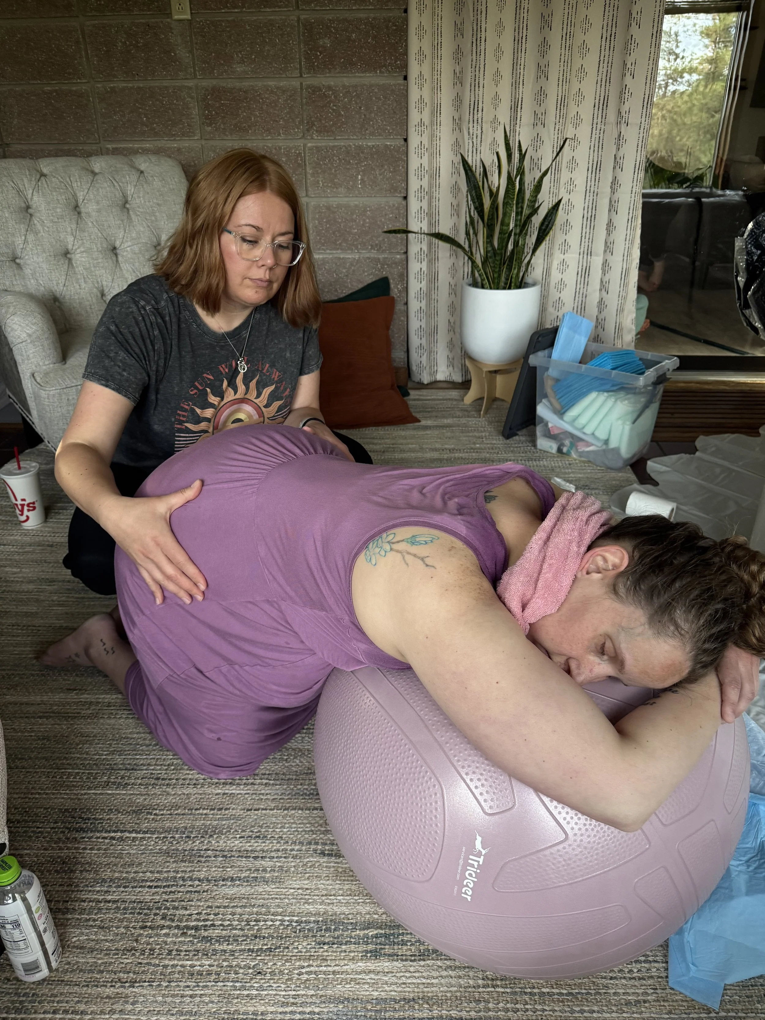 A woman receiving a massage while lying face down on a purple yoga ball in a room with a gray carpet, potted plant, curtains, and an outdoor view through the window.