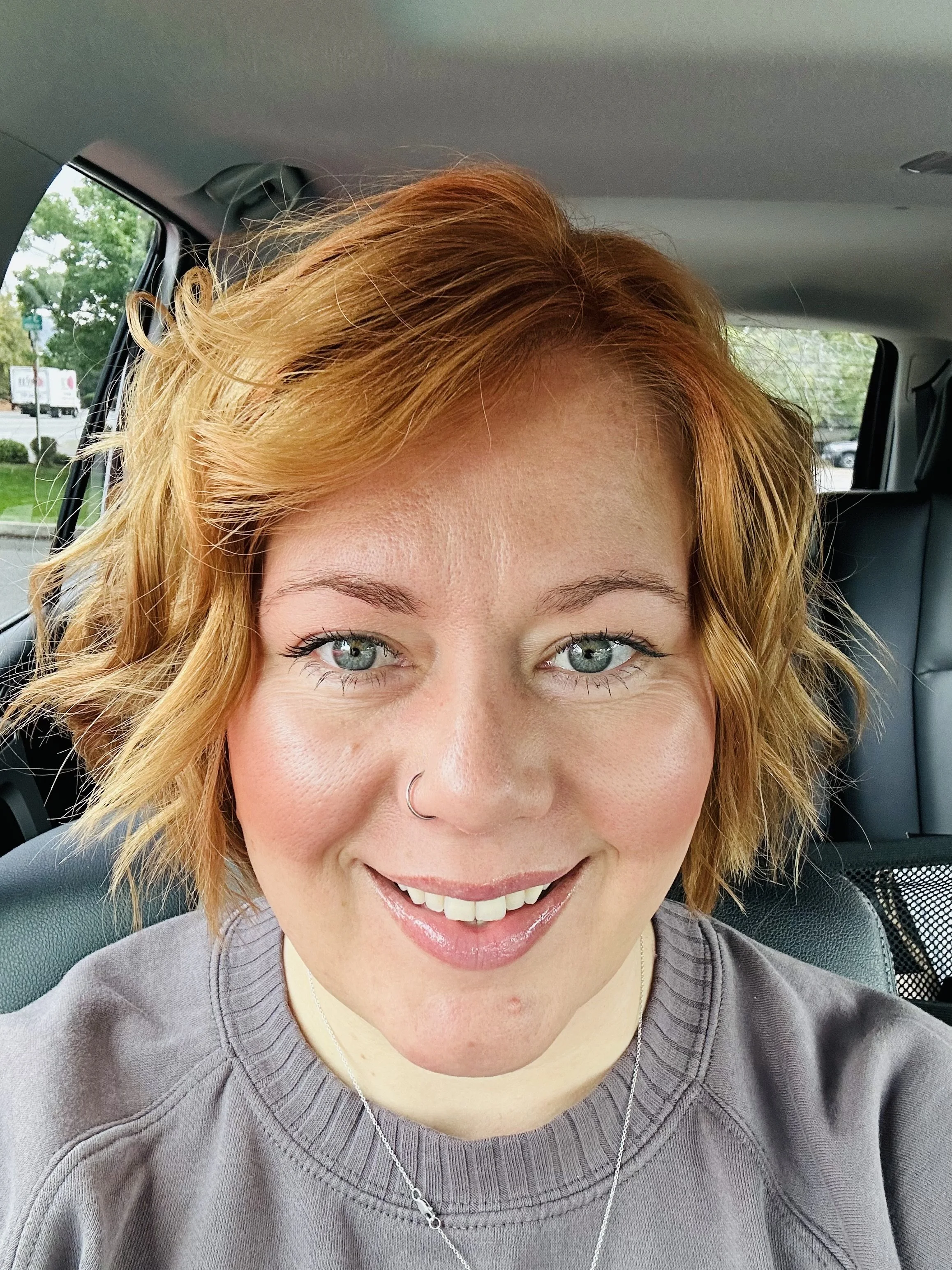 Close-up of a woman with short, wavy, red hair, smiling and wearing a purple sweatshirt and a necklace, sitting in a car.