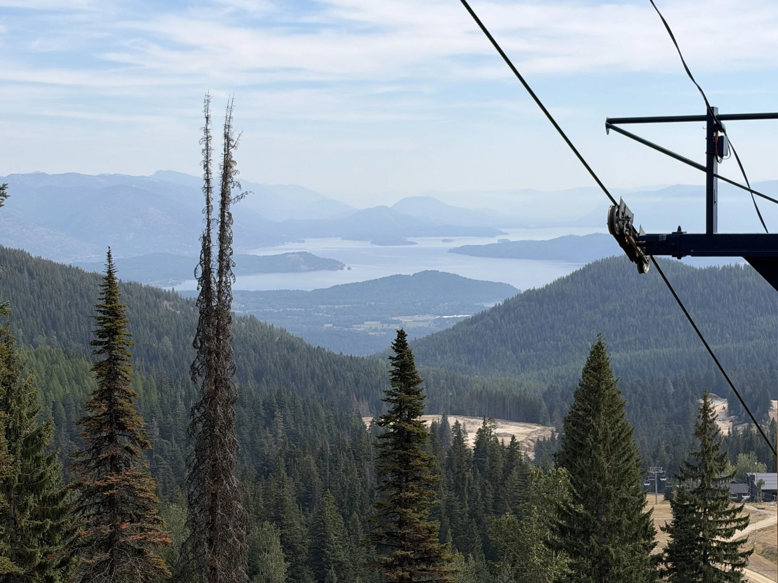 A mountain landscape with dense green pine trees in the foreground, a sky with scattered clouds, and a large body of water and distant mountains in the background. A metal structure on the right side appears to be part of a ski lift or similar feature.