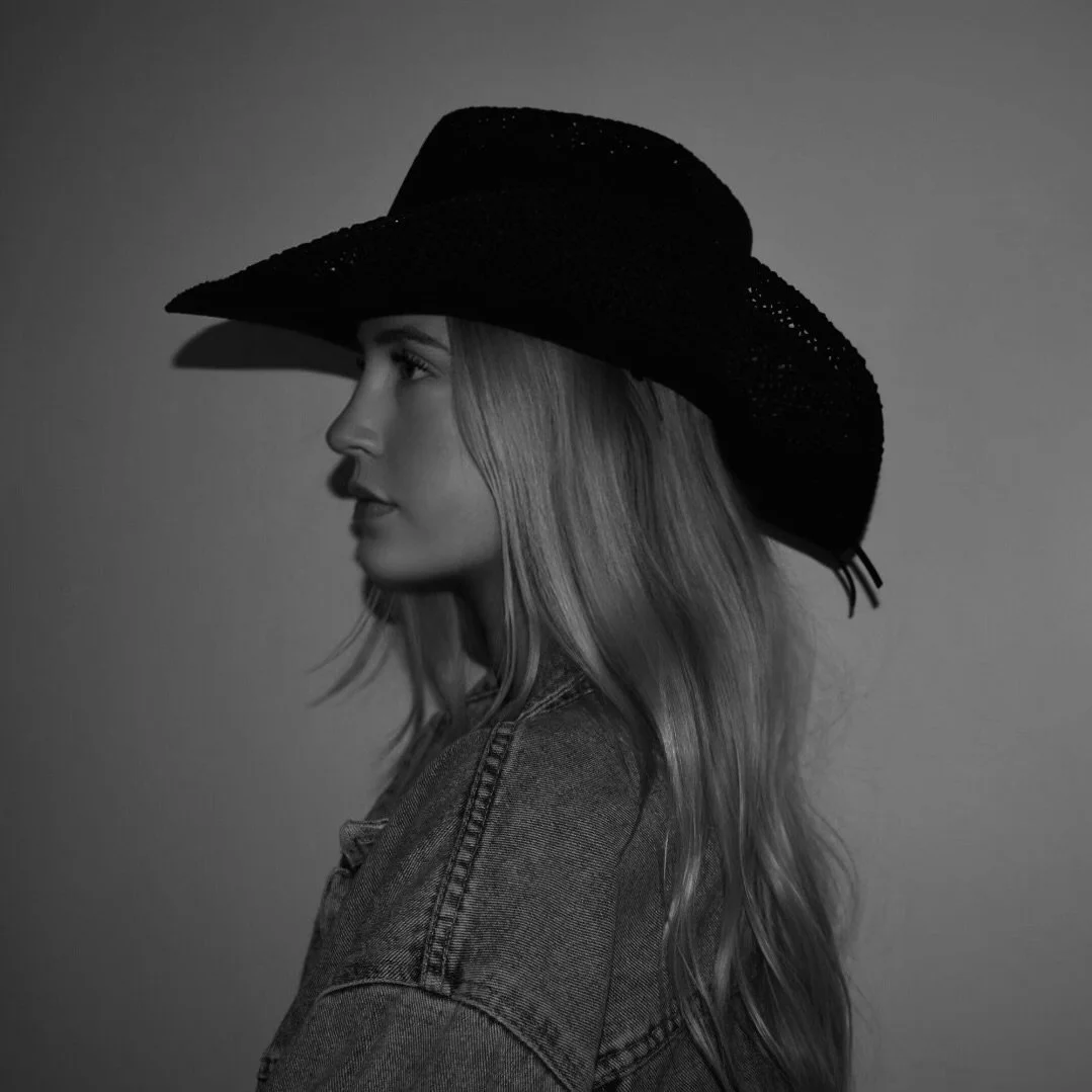 A black and white photo of a woman in profile wearing a wide-brimmed cowboy hat and a denim jacket.