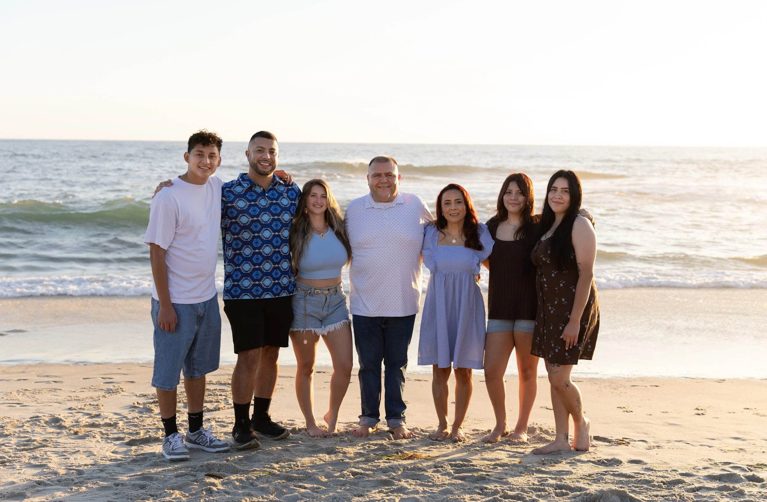 Group of seven friends posing together on a sandy beach at sunset