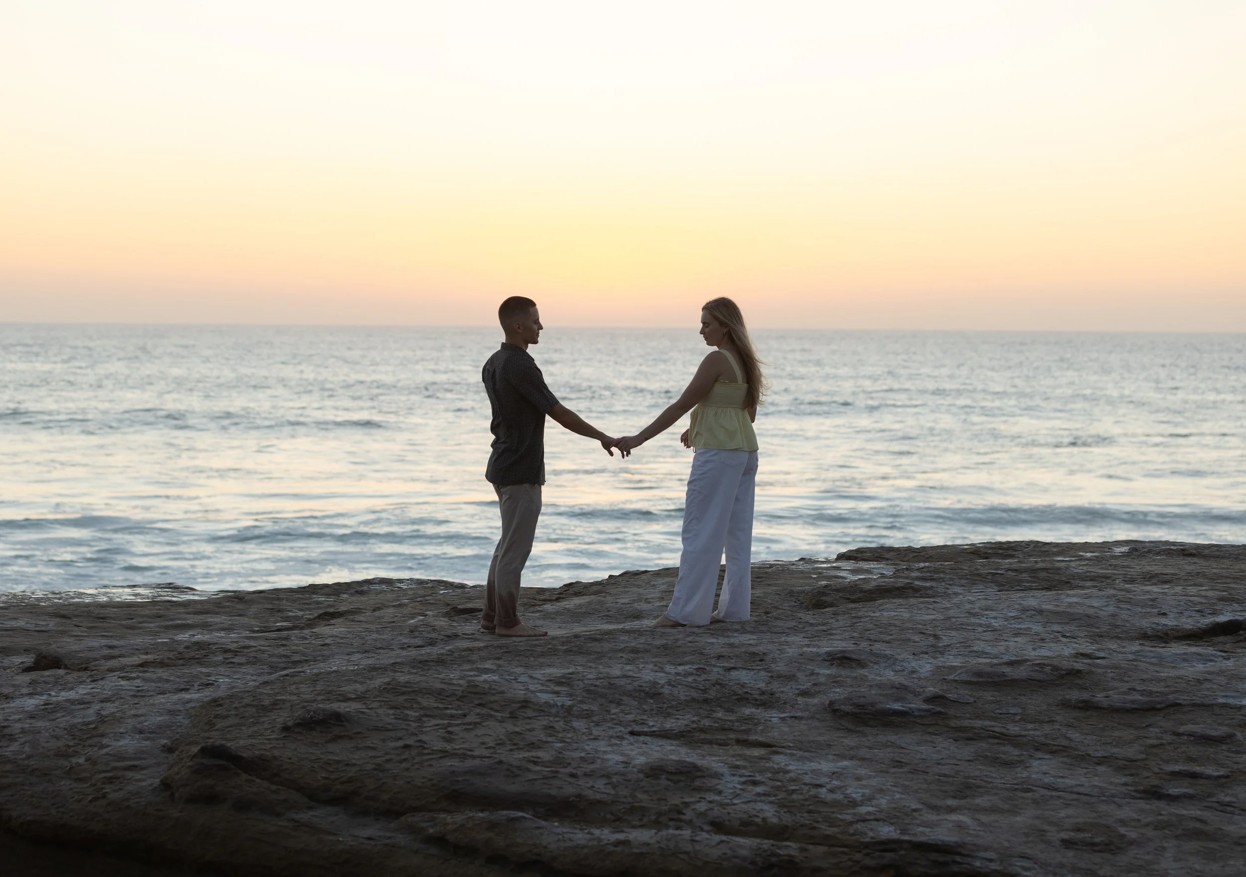 Couple holding hands on rocky shore overlooking the ocean at sunset