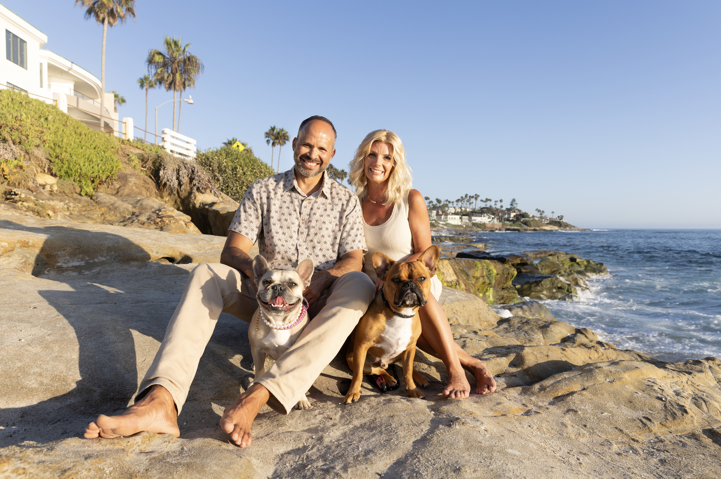 Smiling couple with two French bulldogs sitting on coastal rocks beside the ocean and palm-lined homes
