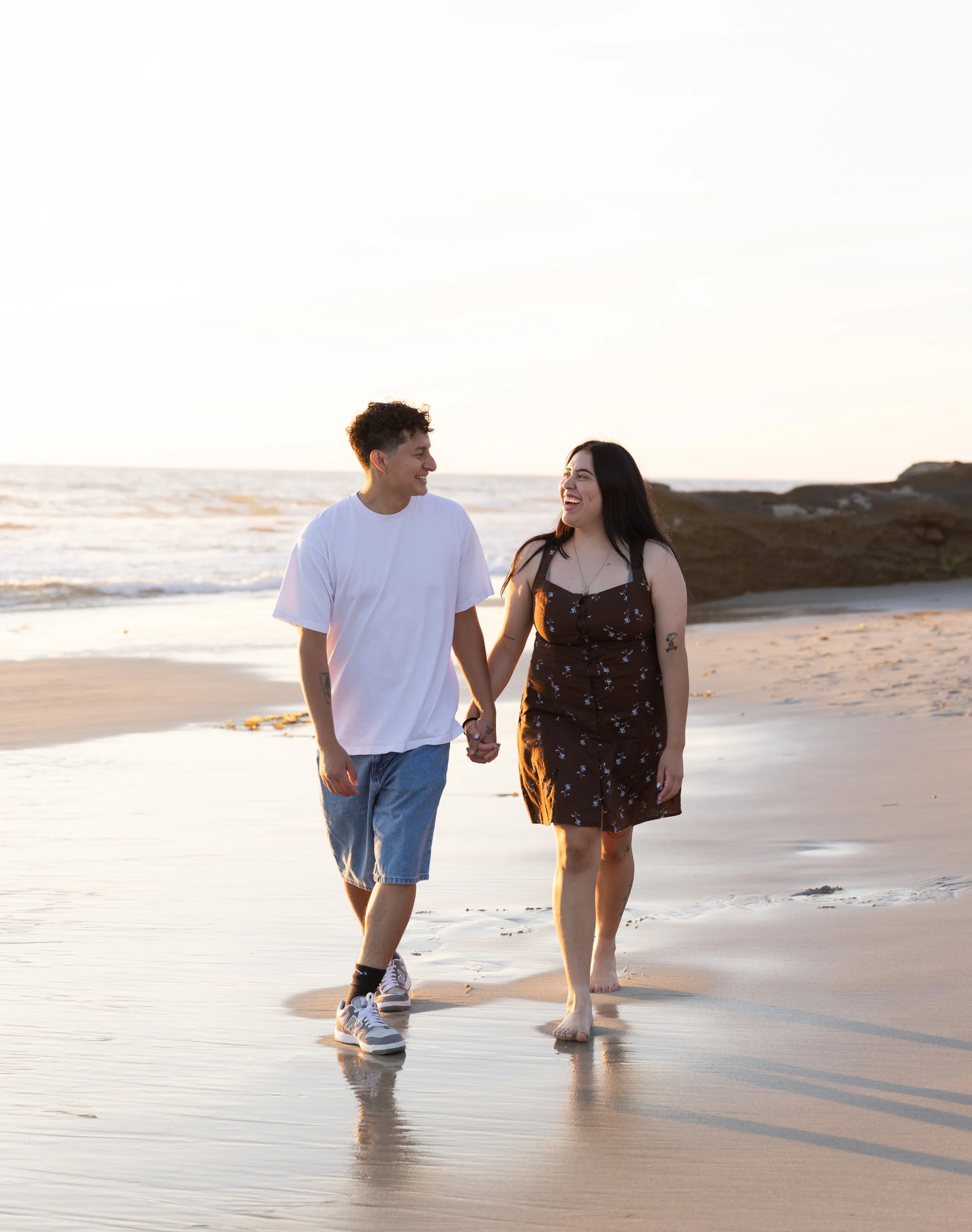 A happy couple holding hands and walking along a beach at sunset, with ocean waves and rocky cliffs behind them.