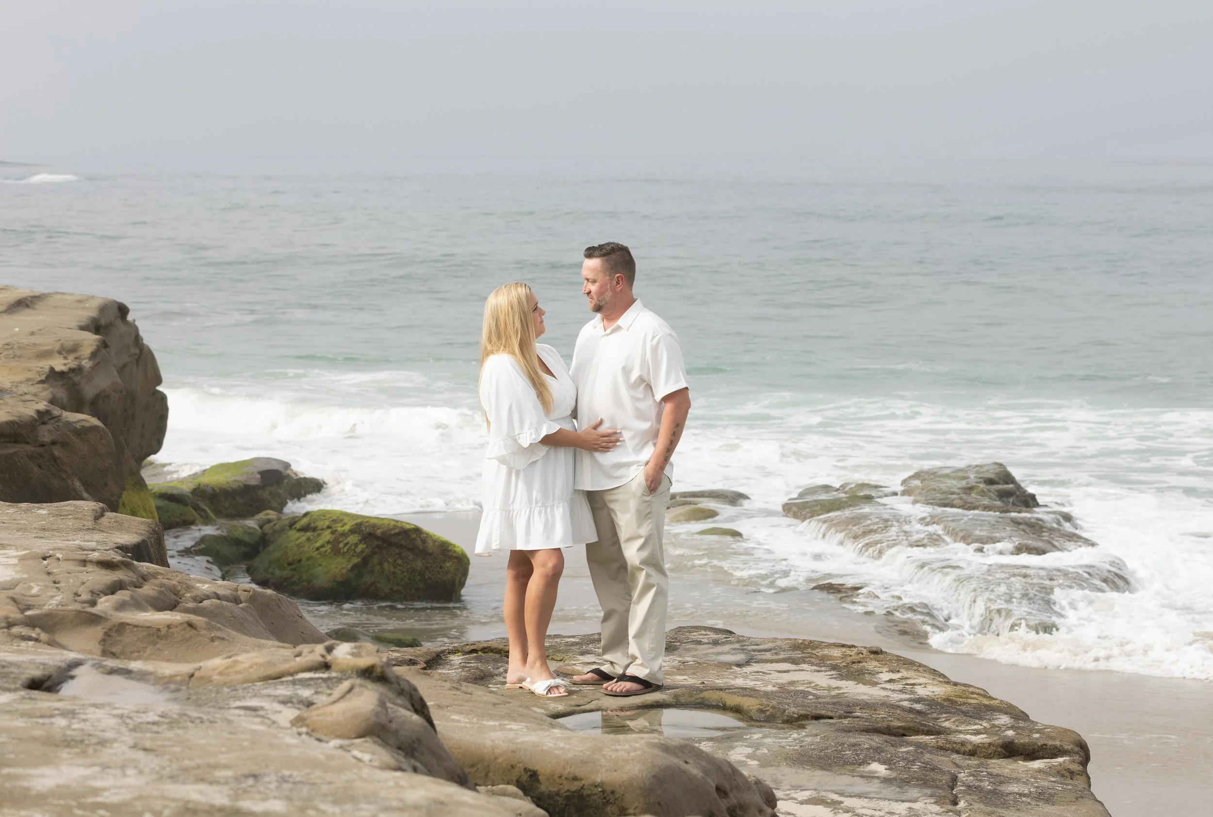 Couple in white clothing standing on rocky sea cliffs beside ocean waves