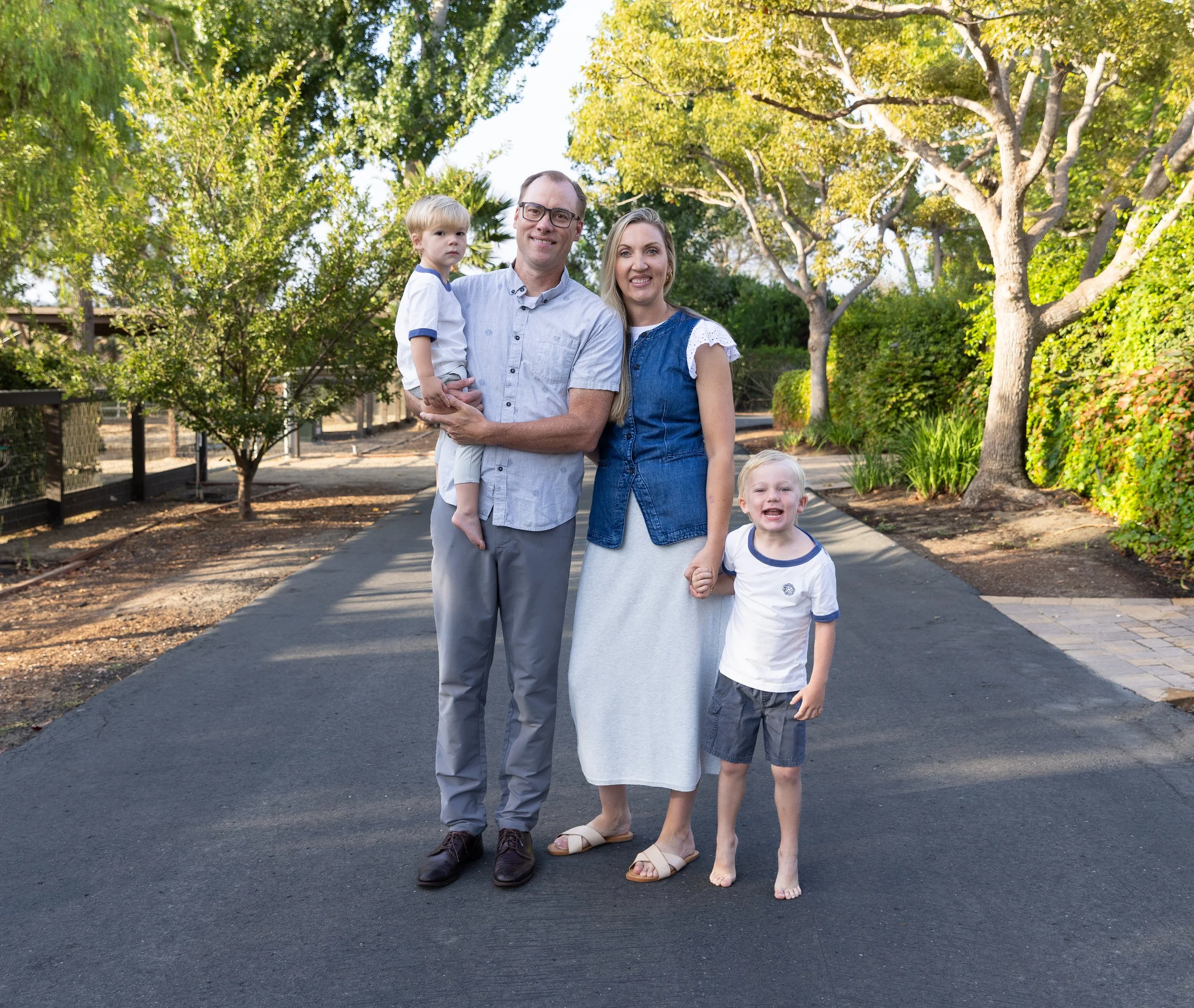 Family of four posing on a tree-lined outdoor walkway, with the father holding a young child and the mother holding another child's hand.