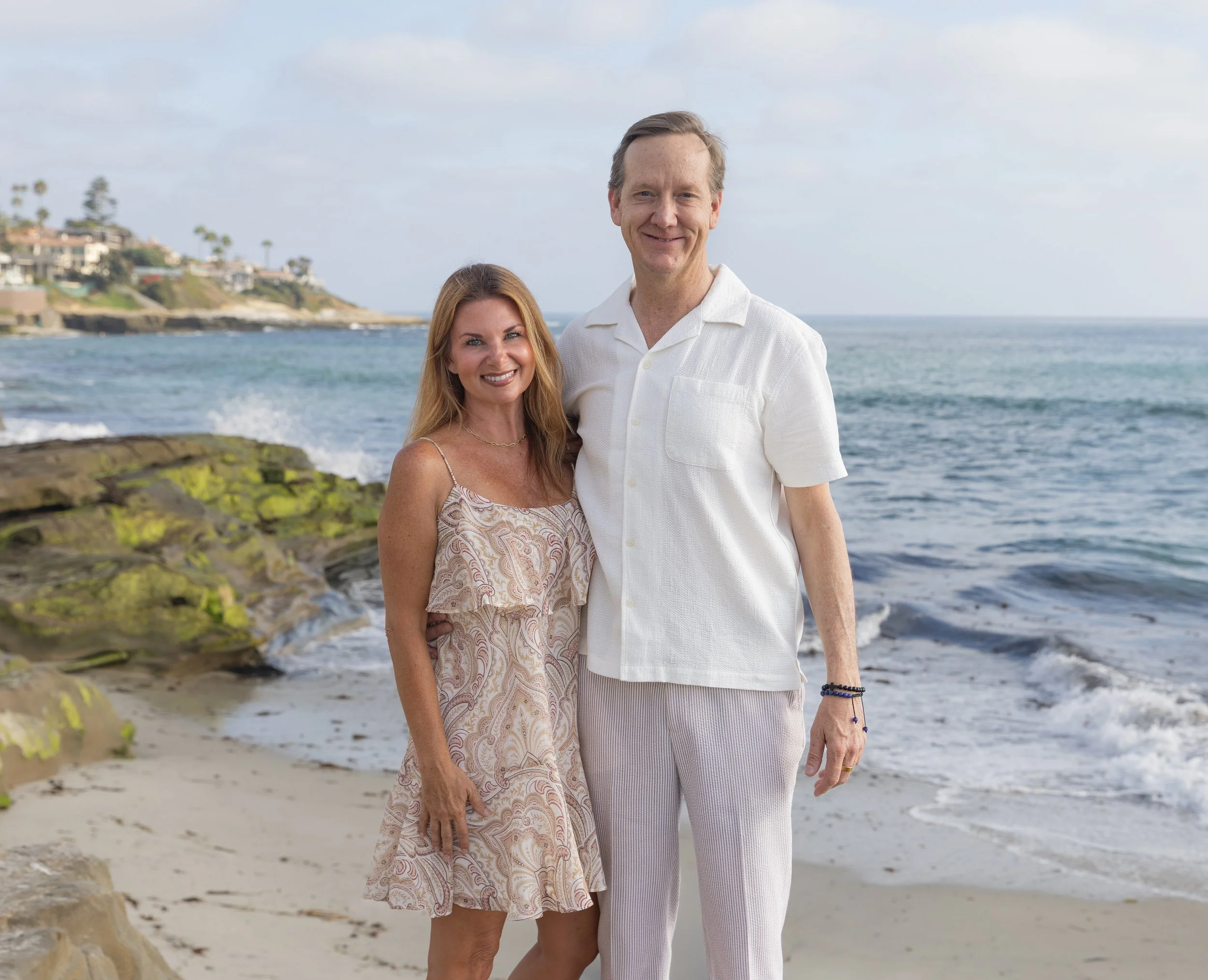 Smiling couple posing on a rocky beach beside the ocean with coastal homes in the background