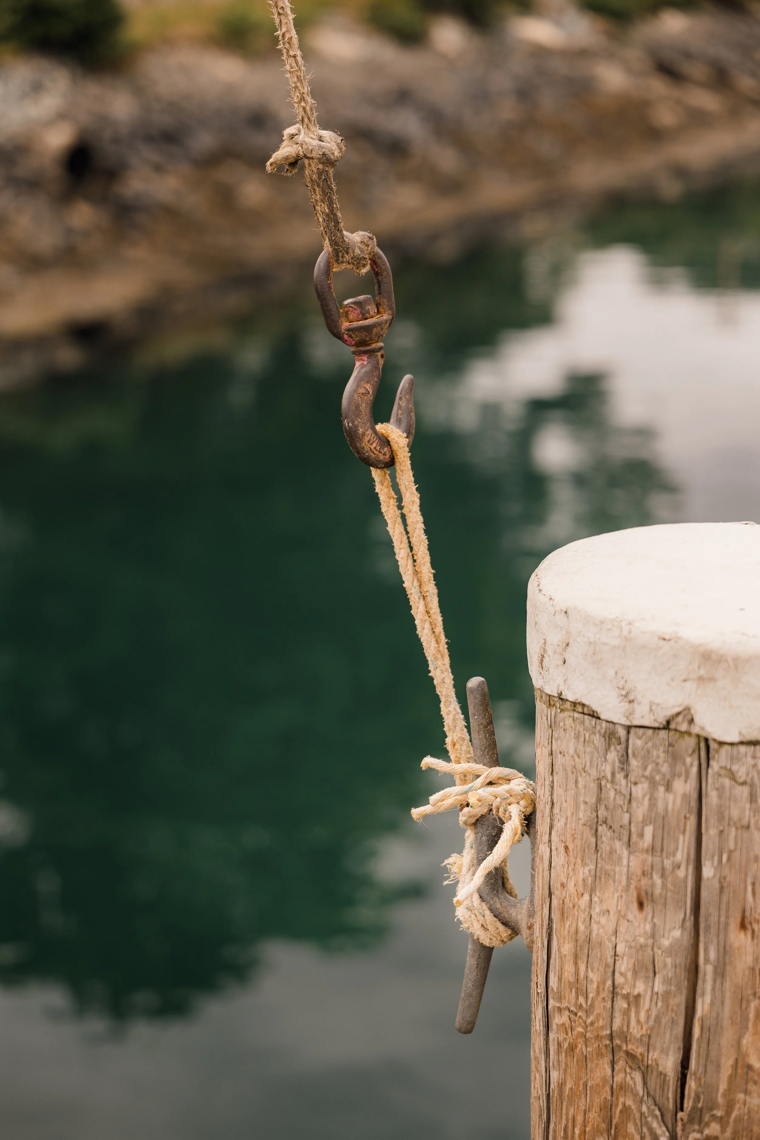 Close-up of a rusty metal hook attached to a weathered wooden post with a frayed rope tied around it, against a blurred body of water.