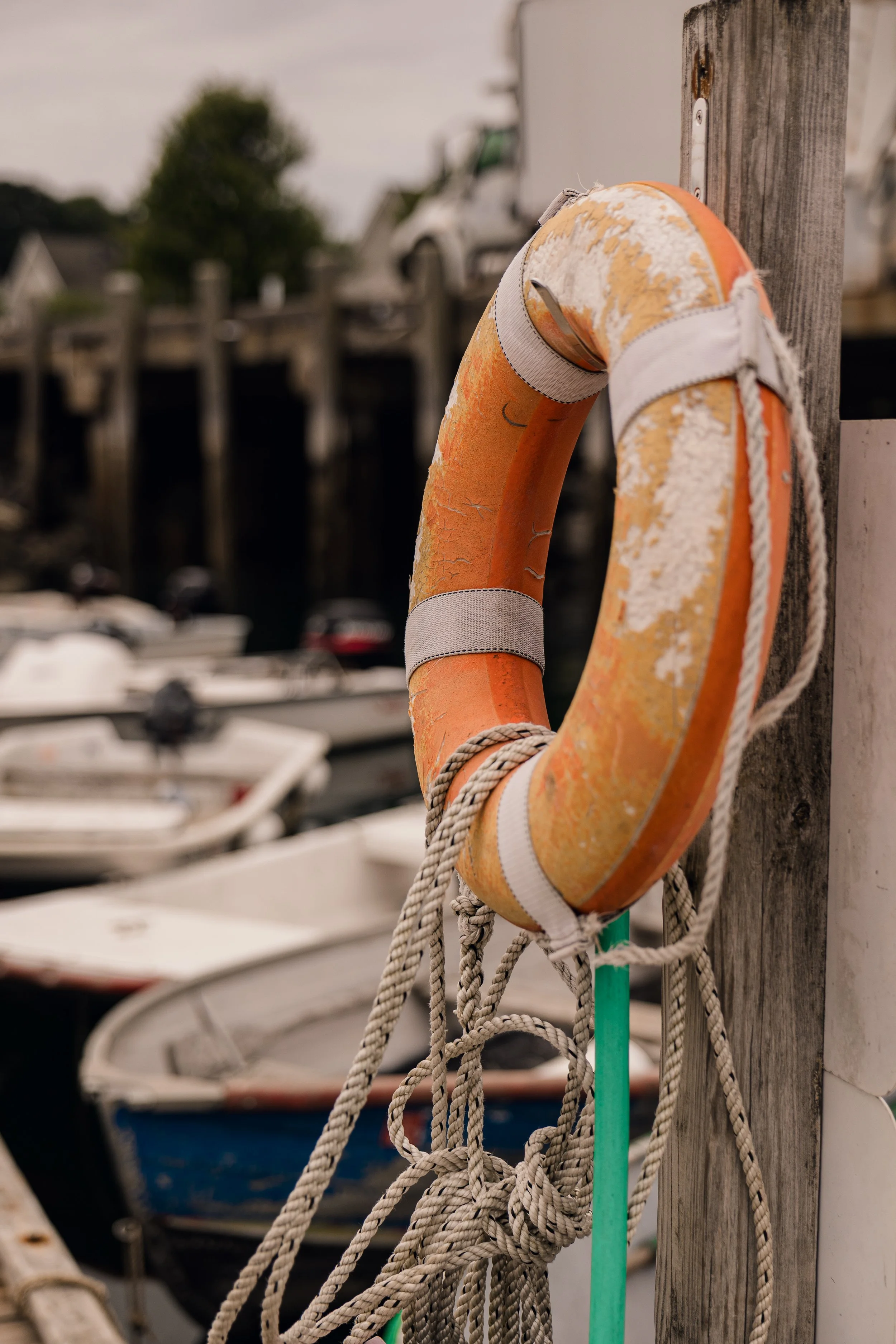A weathered orange and white life preserver ring with frayed rope hanging on a wooden post at a marina, boats and trees blurry in the background.