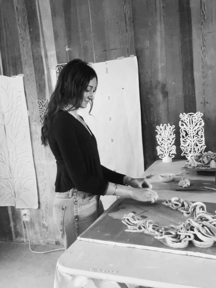 Woman in black top and light jeans arranging rolled and pinched clay on a table and decorative artwork in the background.