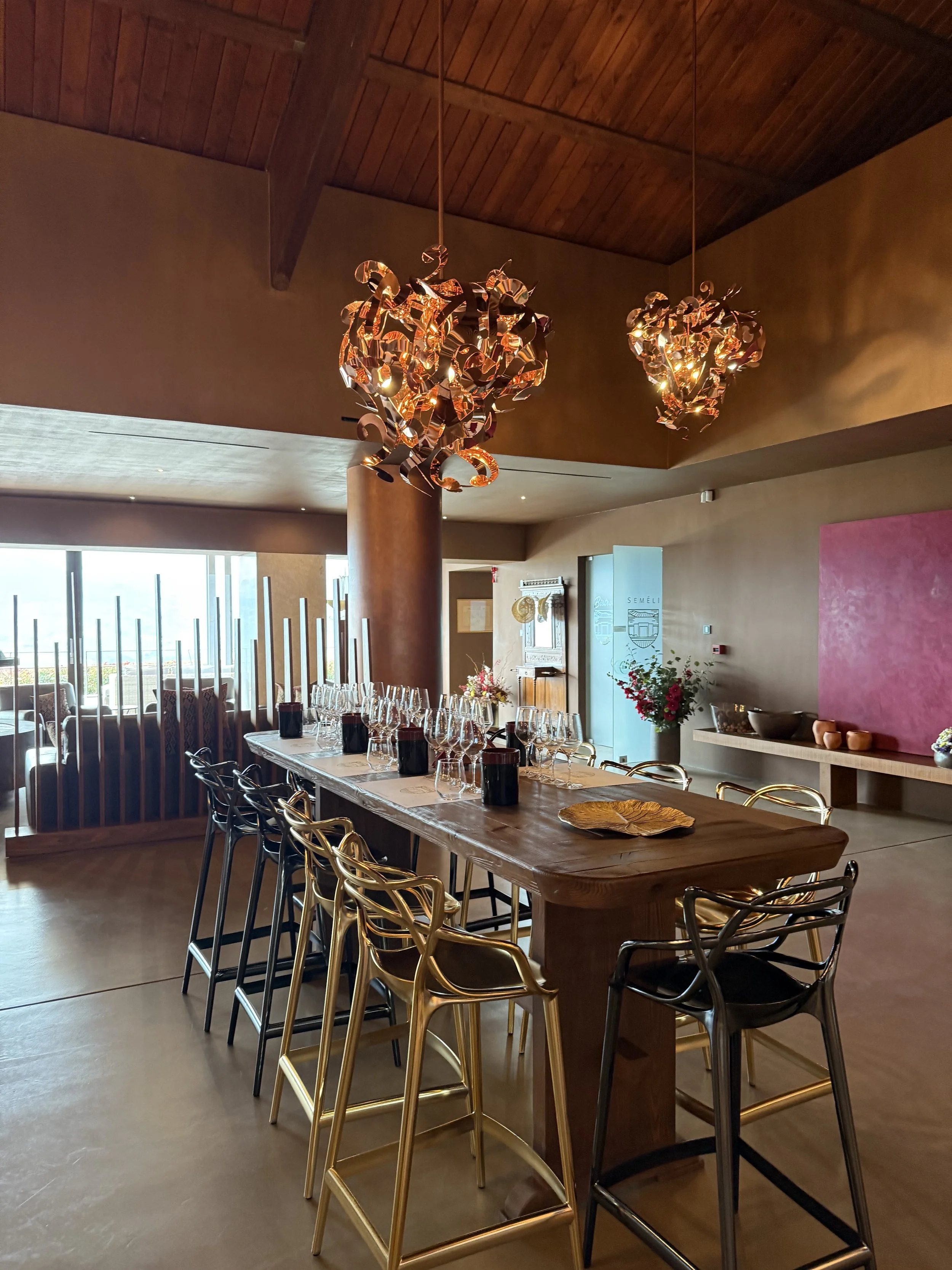 Elegant dining room with a long wooden table set with wine glasses and dark napkins, surrounded by gold and black chairs, featuring modern chandeliers hanging from a wooden ceiling, with large windows letting in natural light.