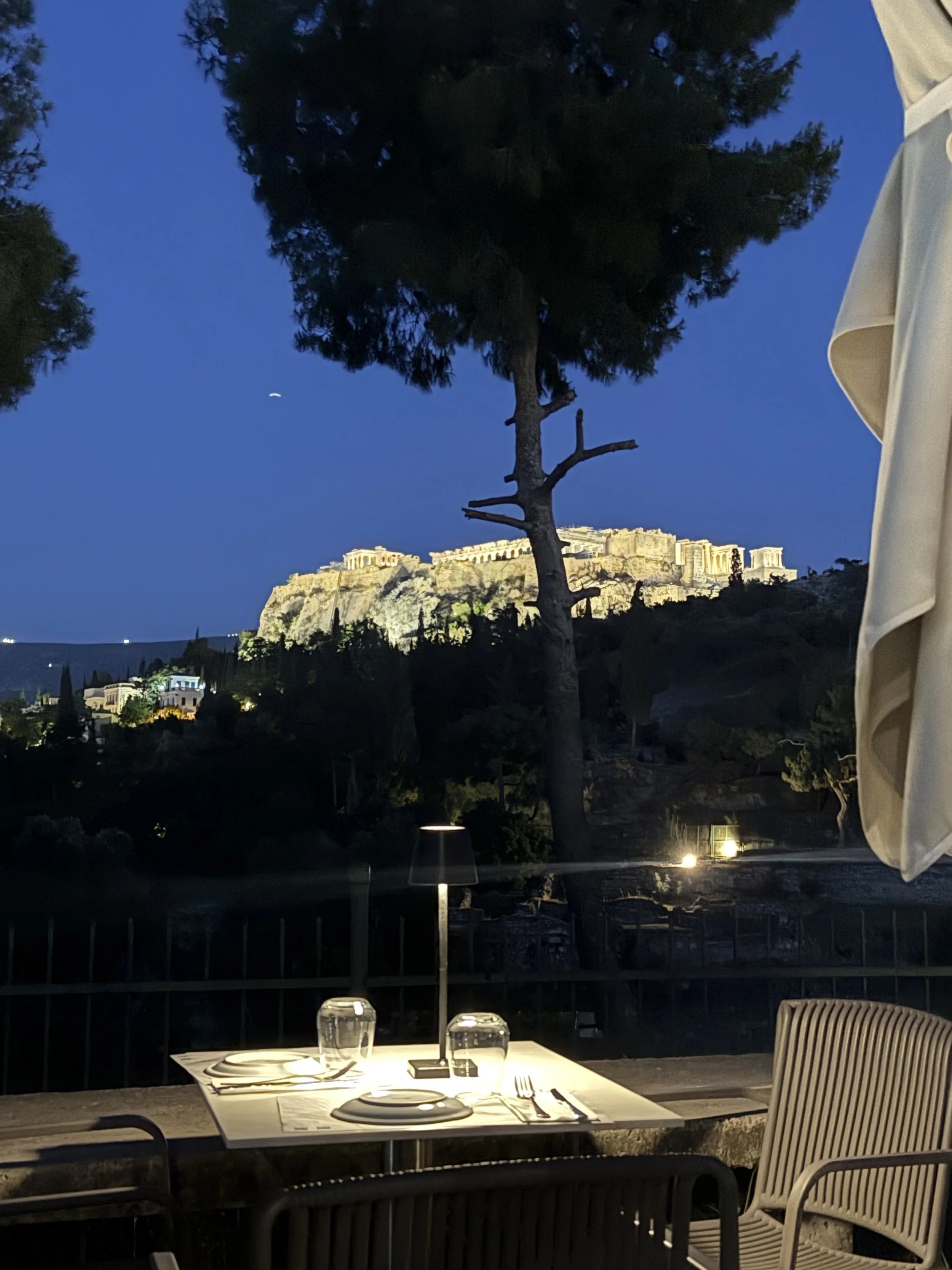 A nighttime outdoor restaurant scene with a table set with plates, glasses, and utensils. In the background, there's a view of the illuminated Acropolis on a hill, with trees and a clear night sky.