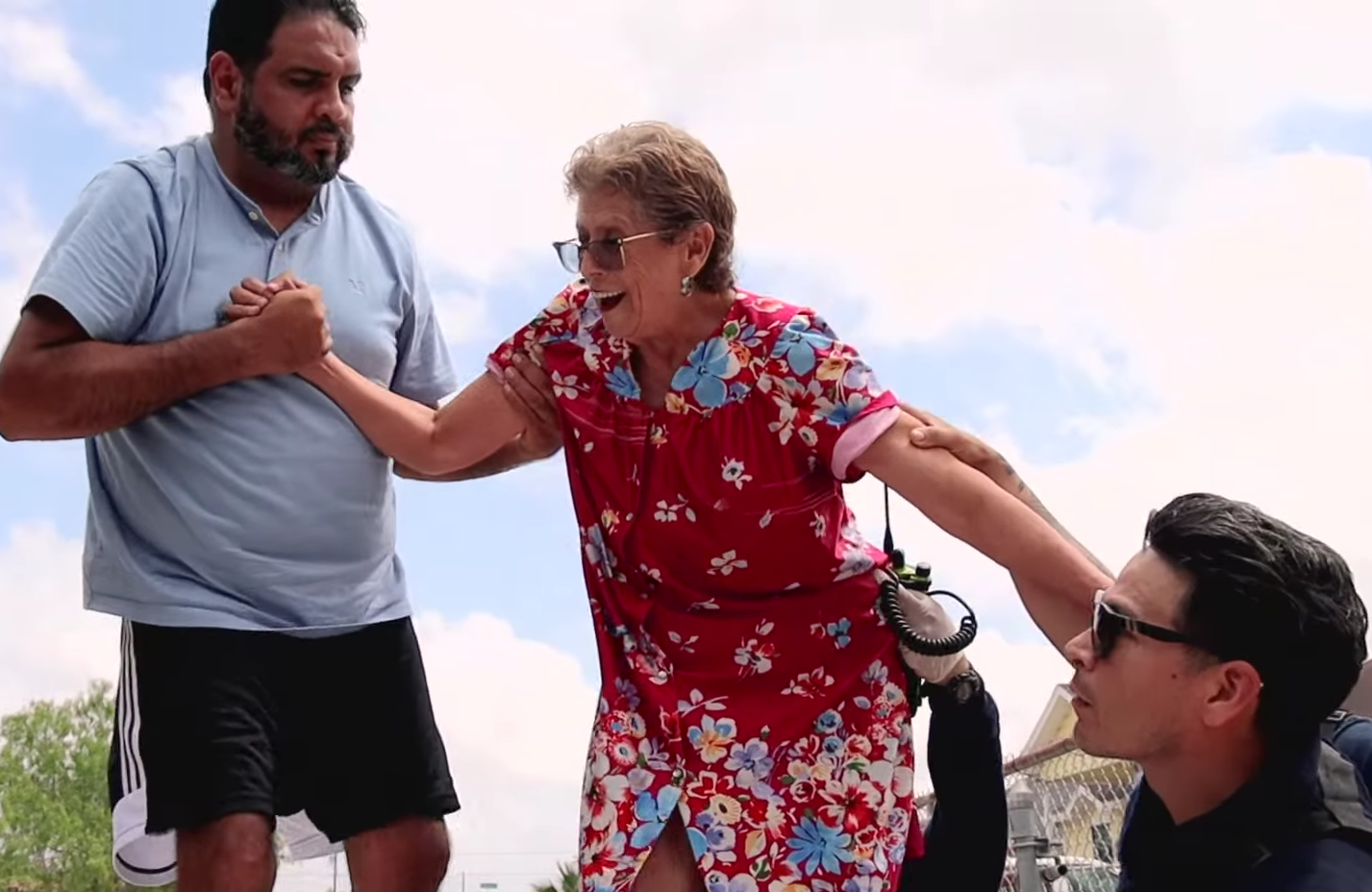 A woman in a floral dress and sunglasses is being assisted by two men — one standing and one sitting — during what appears to be a rescue situation outdoors on a partly cloudy day.