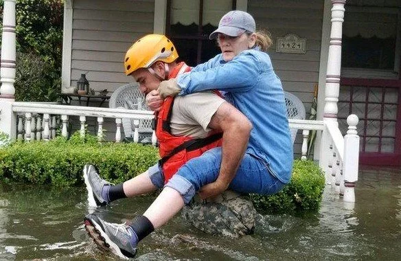 A woman being helped by rescue personnel. She is being carried on his back above the water. He is wearing a yellow helmet
