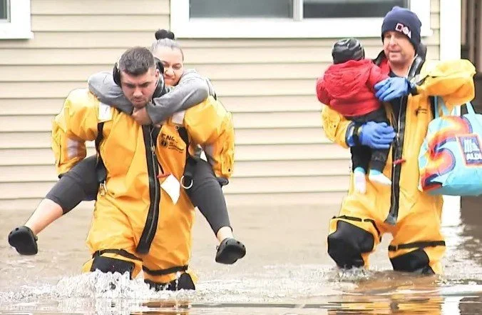 Emergency responders wading through floodwaters, helping children in life jackets.
