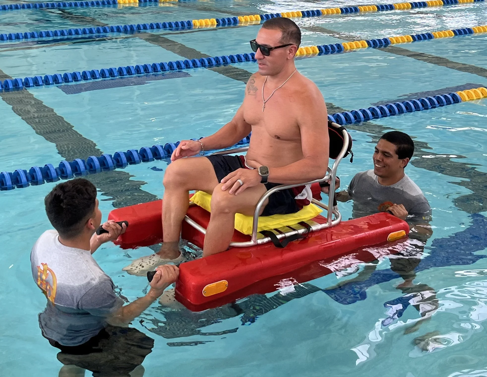 A shirtless man wearing sunglasses and a cross necklace sits on a rescue float in a swimming pool, surrounded by several fire fighters training on a rescue rider device.