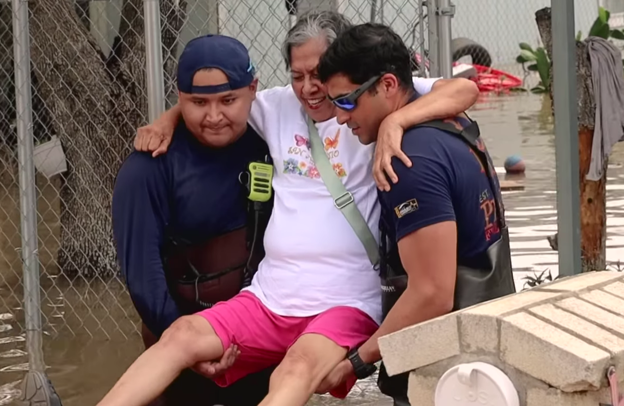 Three men are rescuing a person from floodwaters. The person being rescued is sitting on the lap of one rescuer and appears to be comforted by the others. Flooded area with water, debris, and fencing is visible in the background.