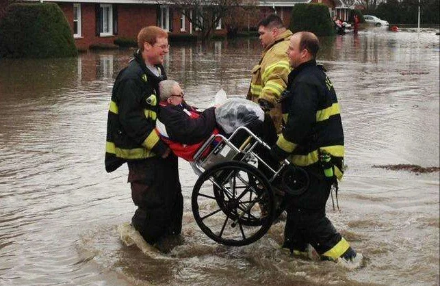 Three firefighters rescuing a person in a wheelchair from a flooded street
