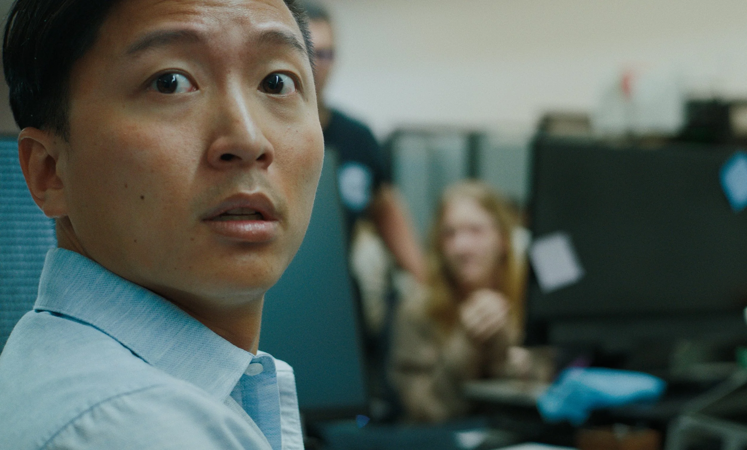 Close-up of a young Asian man in a blue collared shirt looking at the camera, with a blurred background showing a woman working at a computer in a busy office.