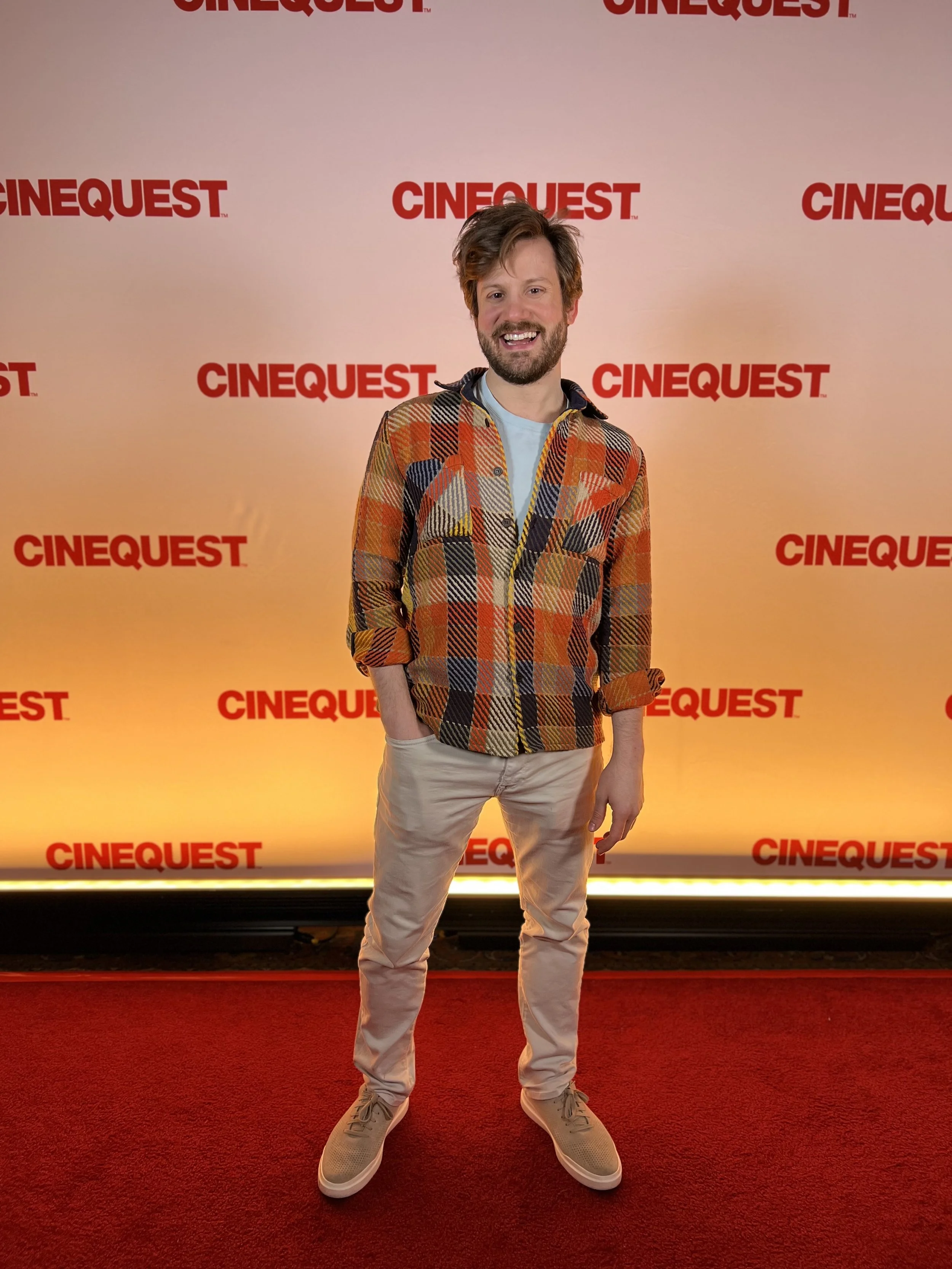 A man with brown hair and a beard is smiling at the camera. He is wearing a colorful, patterned jacket, light-colored pants, and beige sneakers. He is standing on a red carpet in front of a yellow background with the red text "CINEQUEST" repeatedly printed on it.