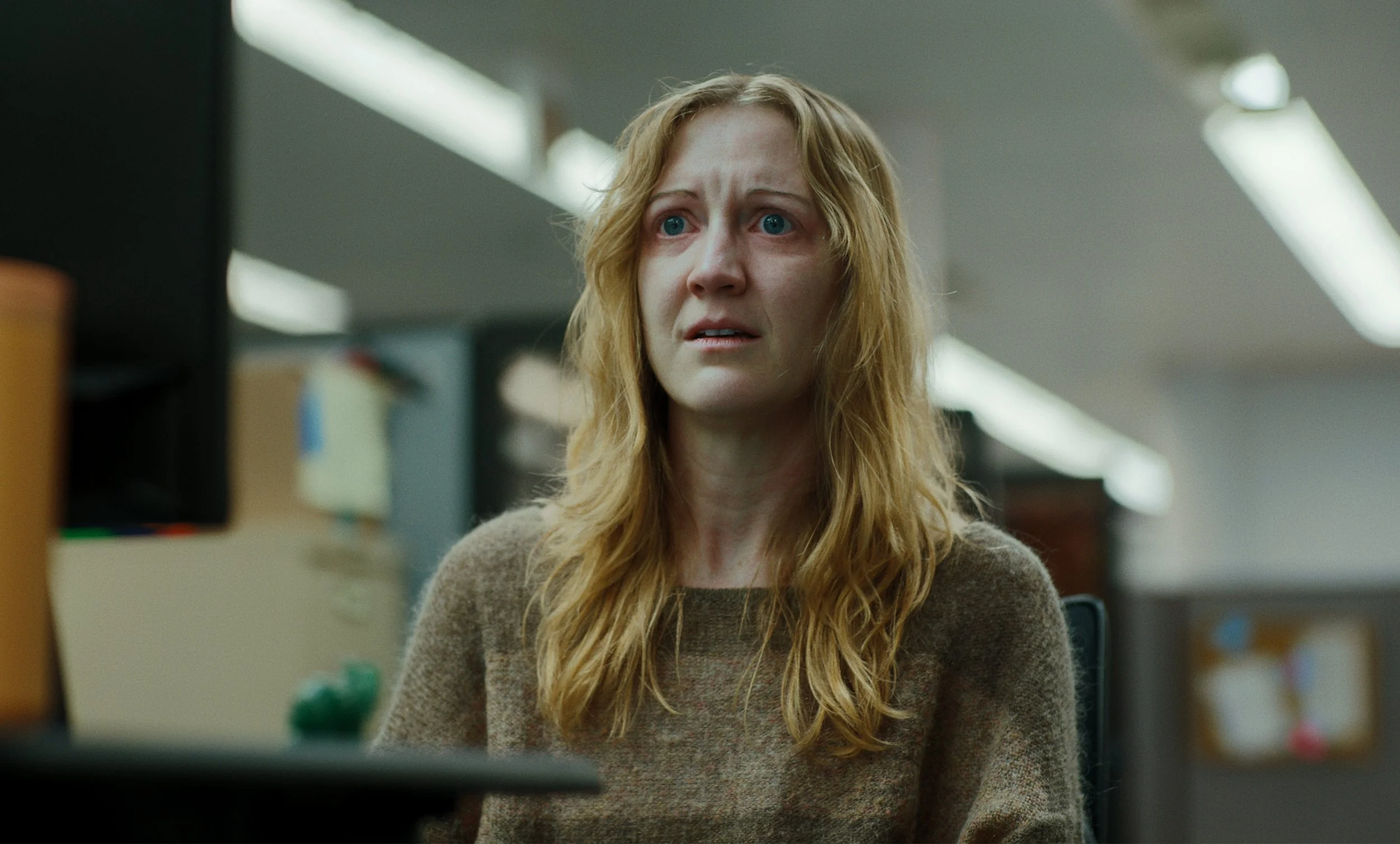 A woman with red hair and blue eyes looking at a computer screen with a concerned expression in an office environment.
