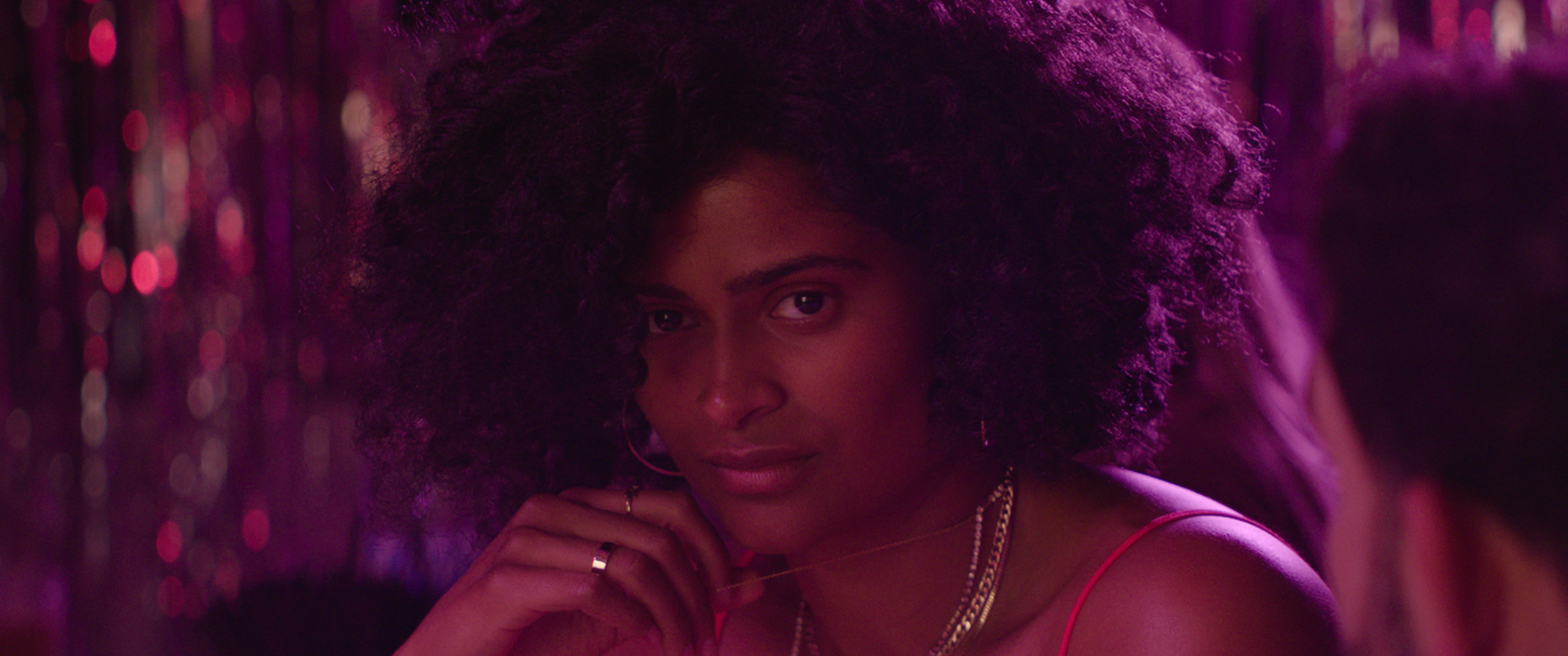 A young woman with curly hair and hoop earrings looking directly at the camera, with a pink and purple lit background.