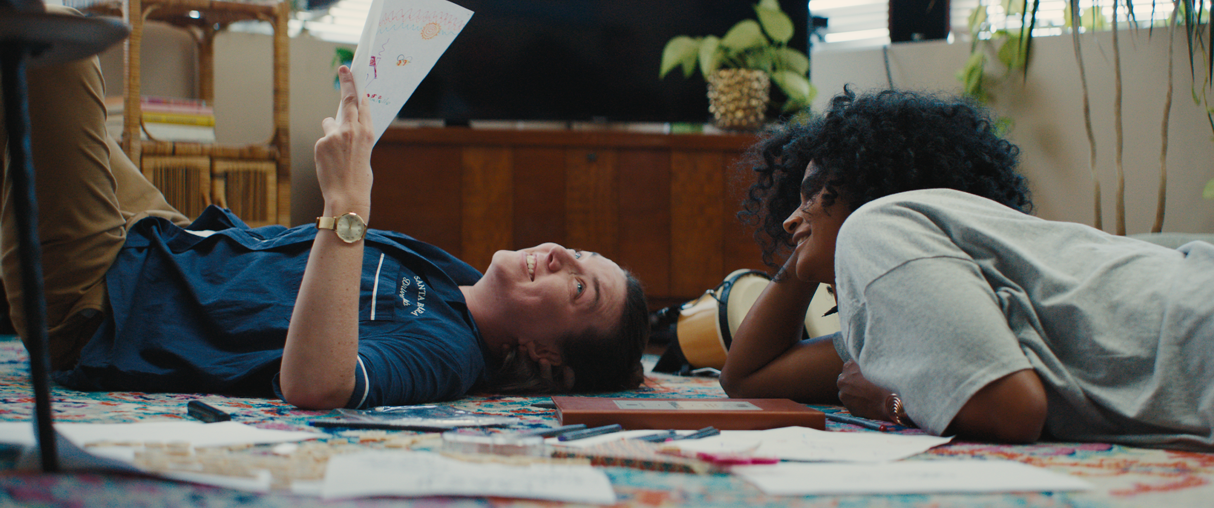 Two women are lying on a colorful rug in a cozy room, engaging in a lively conversation. One woman is on her back, holding a piece of paper, while the other woman is leaning on her elbows, smiling. There are books, a small box, and musical instruments around them, with plants and a TV in the background, creating a warm, inviting atmosphere.