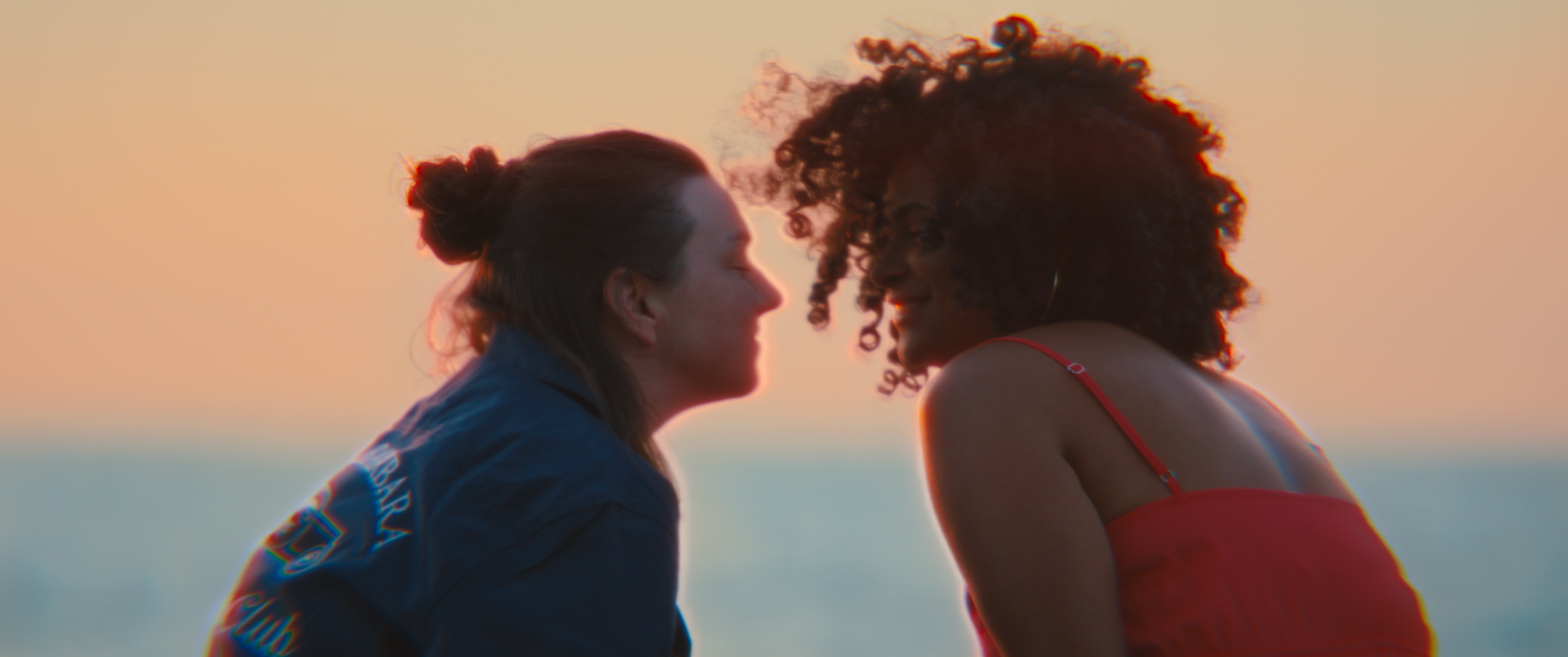Two women with faces close together, smiling, outdoors during sunset, one with dark curly hair wearing a red top and the other with dark straight hair in a bun wearing a blue jacket.