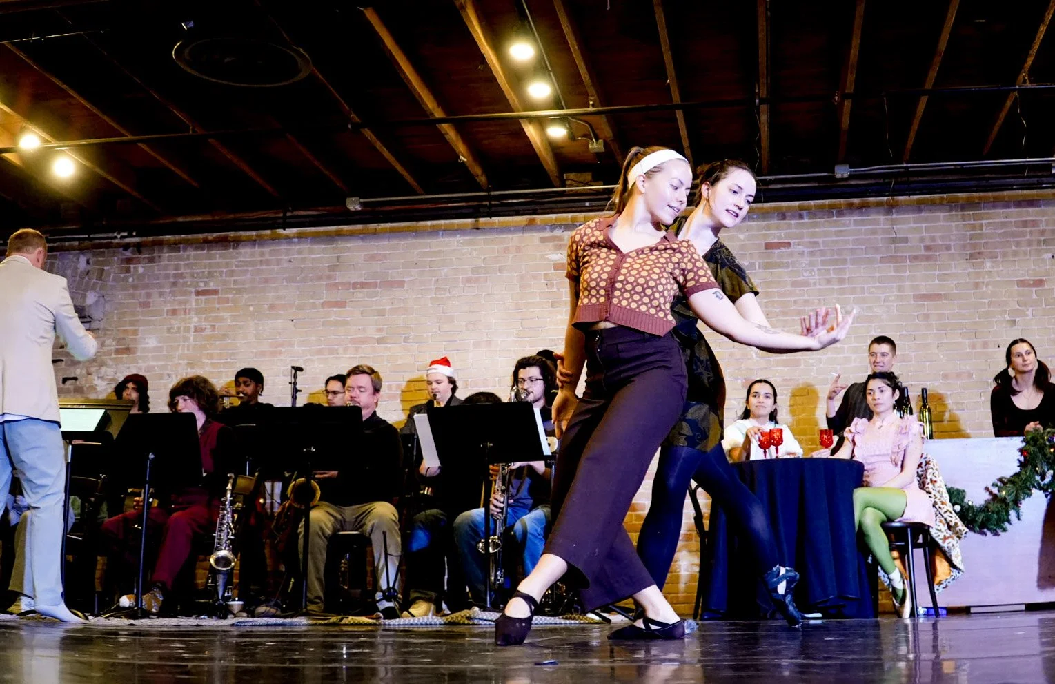 Two women dancing in front of an orchestra on a stage decorated for Christmas, with a brick wall background.
