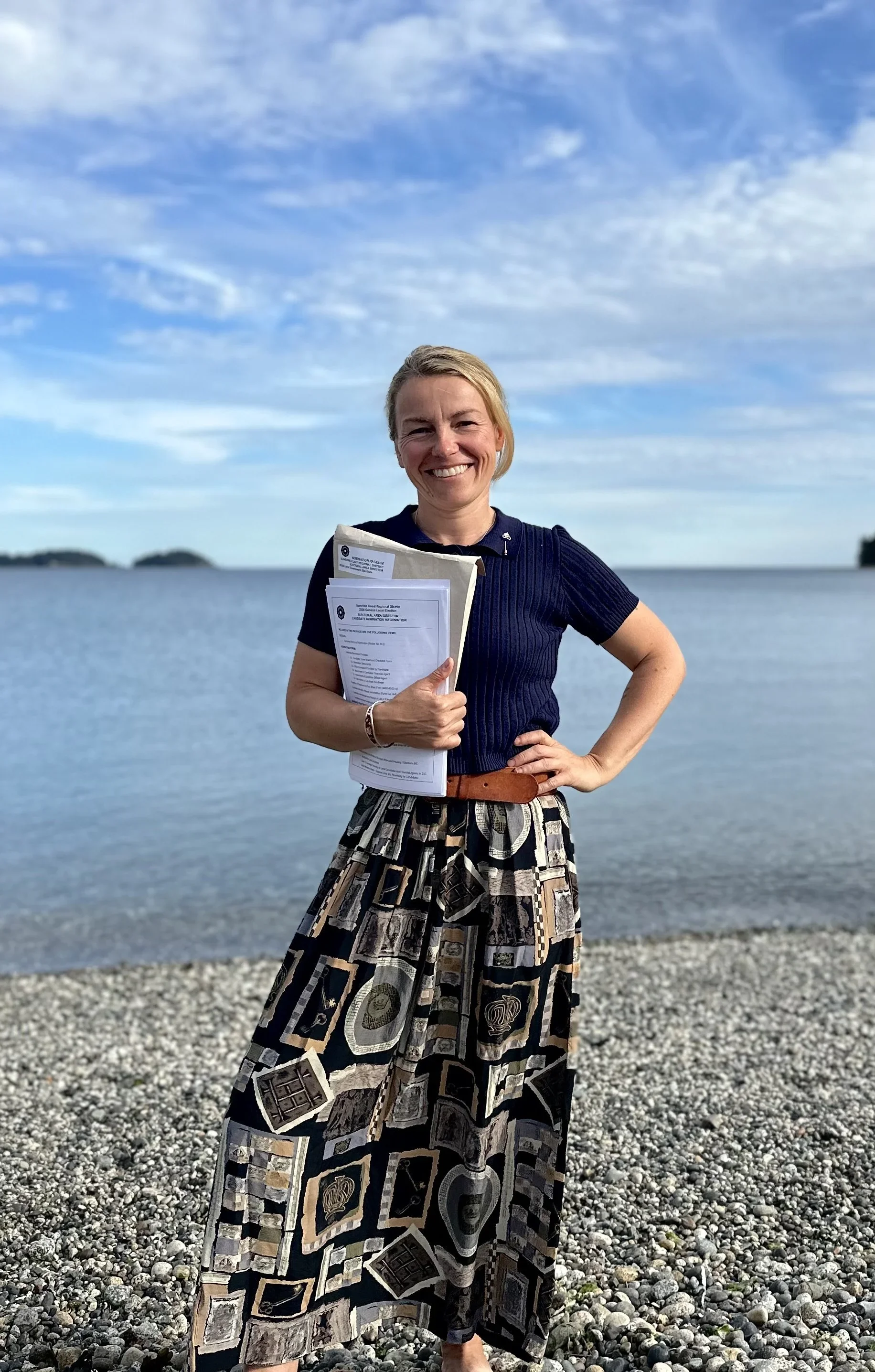 A photograph of Justine Gabias standing on a rocky beach near water, holding papers, wearing a patterned skirt and a dark top, with a partly cloudy sky in the background.