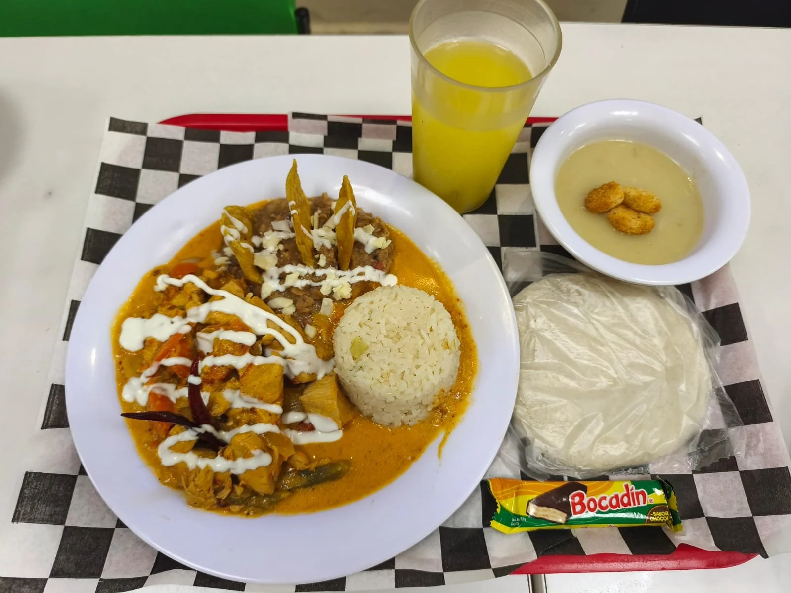Plato con arroz, chili con carne, totopos, crema, y verduras en salsa, acompañado de una bebida de limón, sopa con unas galletas, pan, y chocolate Bocadín.