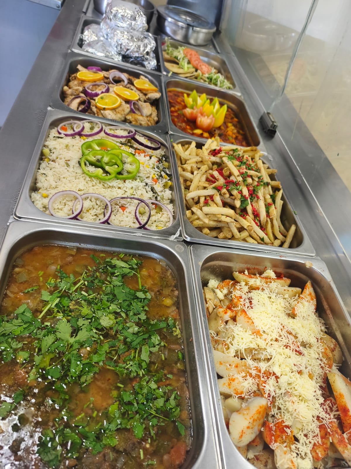 Variedad de comida mexicana en contenedores de acero inoxidable, incluyendo arroz con verduras, carne en salsa, ensalada con limón y cebolla, espaguetis con salsa y queso, y otros platillos tropicales.