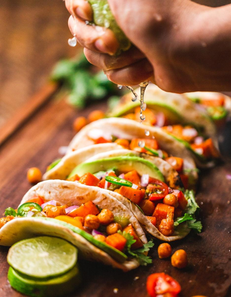 Mano exprimiendo limón sobre tacos con ingredientes variados, incluyendo garbanzos, tomate, cebolla, y cilantro, sobre una tabla de madera.