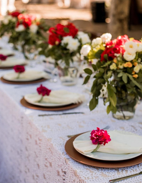 Mesa decorada con platos, cubiertos, servilletas y centros de flores en jarrones con flores rojas, blancas y rosadas.