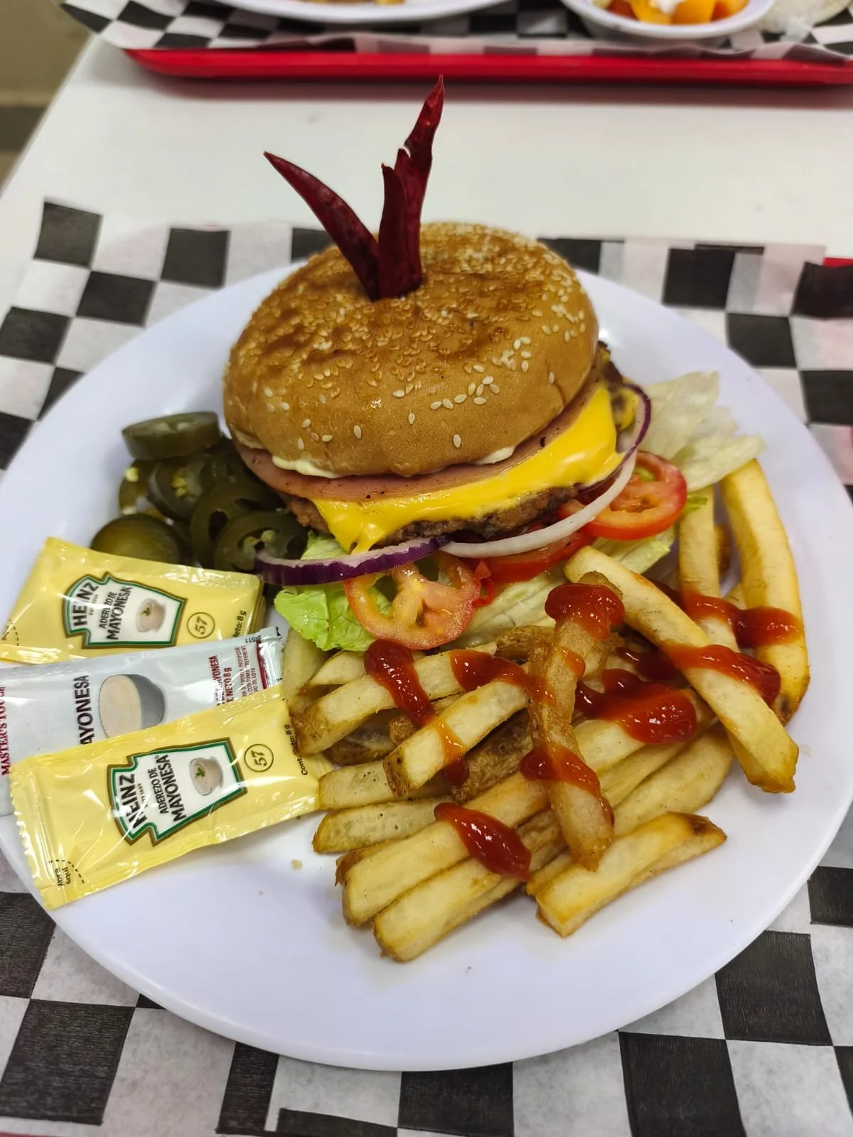 Hamburguesa con papas fritas, ensalada, pepinillos, salsa de tomate y mayonesa, en un plato blanco. La hamburguesa lleva pan con sésamo, carne, queso, jamón, huevo, cebolla, tomate y lechuga. Hay sachets de mayonesa en el plato.