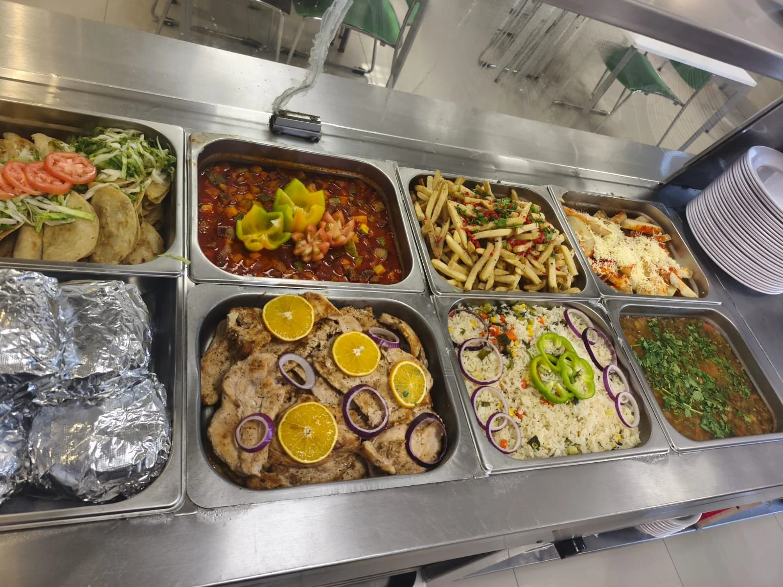 Variedad de platillos mexicanos en una estación de comida, incluyendo pollo con rodajas de naranja, diferentes tipos de pasta, arroz y guiso con cilantro, ensalada y tacos con verduras.