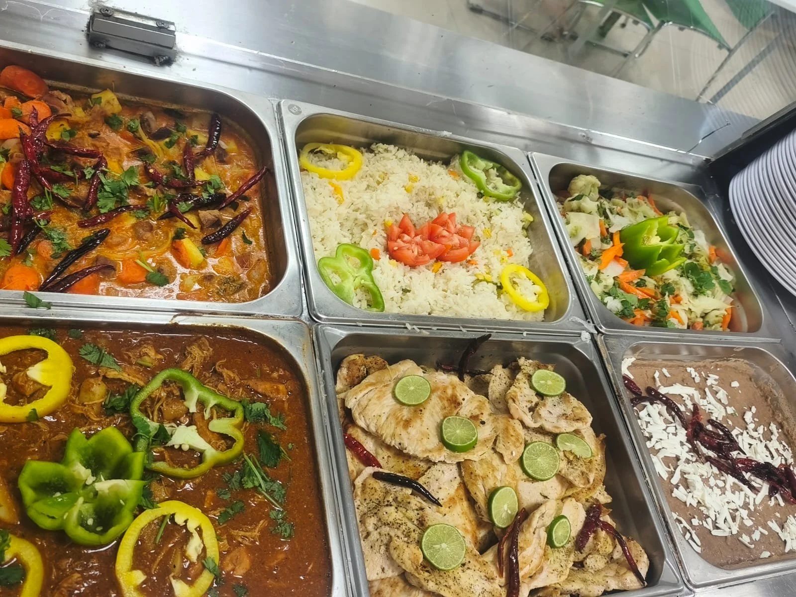 Varios recipientes de acero inoxidable con comida típica mexicana, incluyendo arroz con vegetales, pollo con rodajas de lima, y diferentes tipos de guisos y ensaladas.