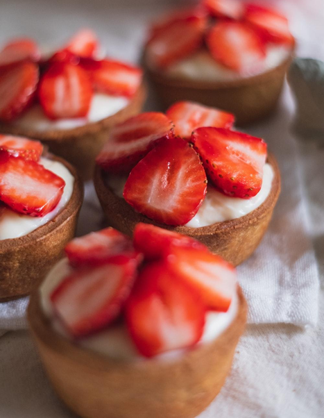 Cuatro pequeñas tazas de postre con base de galleta, crema y fresas en rodajas, sobre un paño de tela beige.